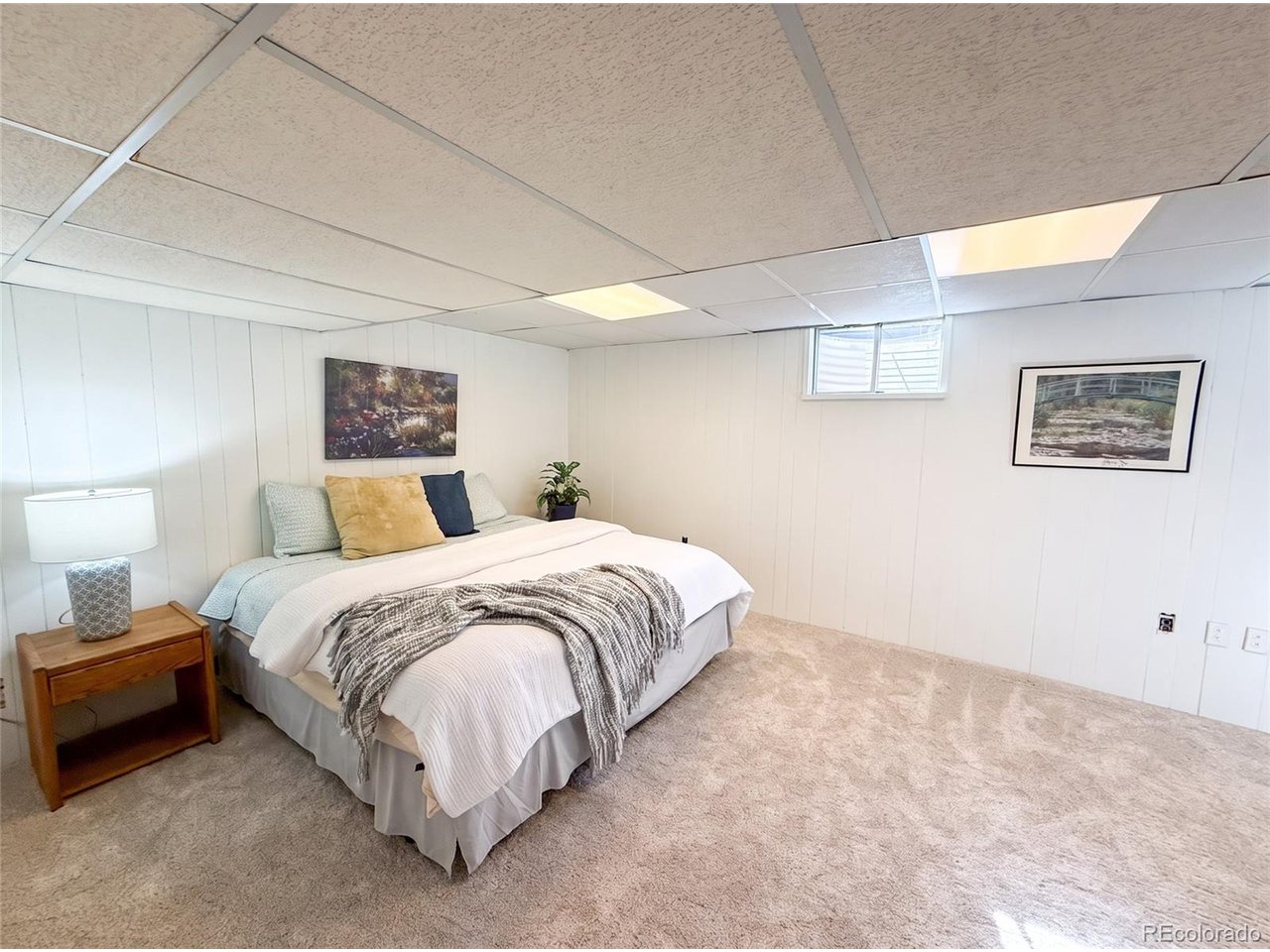Furnished basement bedroom with a white bed, patterned throw, bedside table, and small high window.