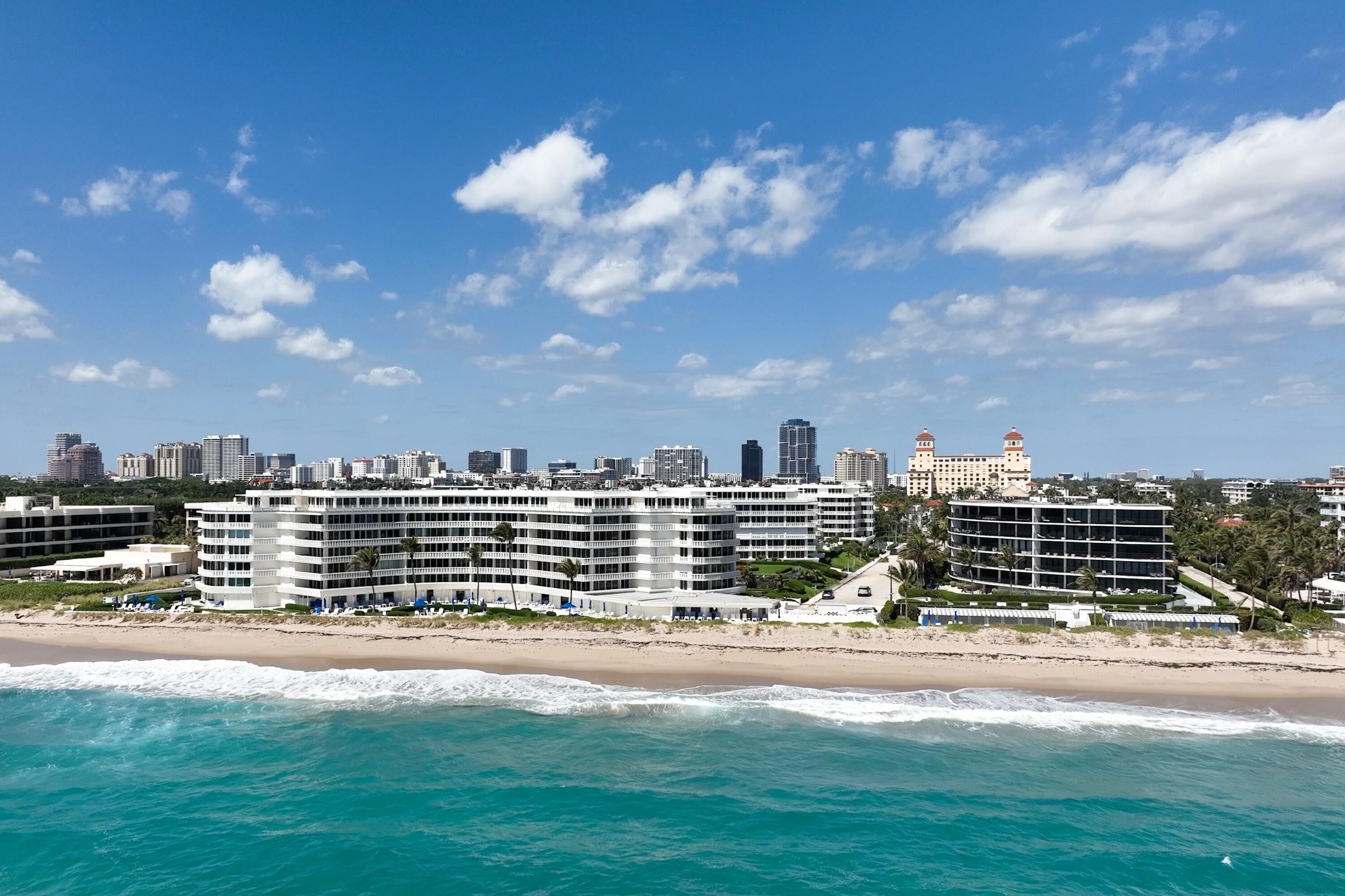 Beachfront condominium buildings along a sandy shore, with turquoise water and a distant city skyline.