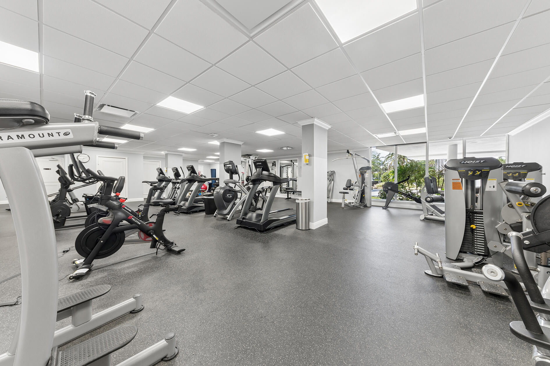 Fitness center with cardio and weight machines beside windows overlooking tropical greenery.