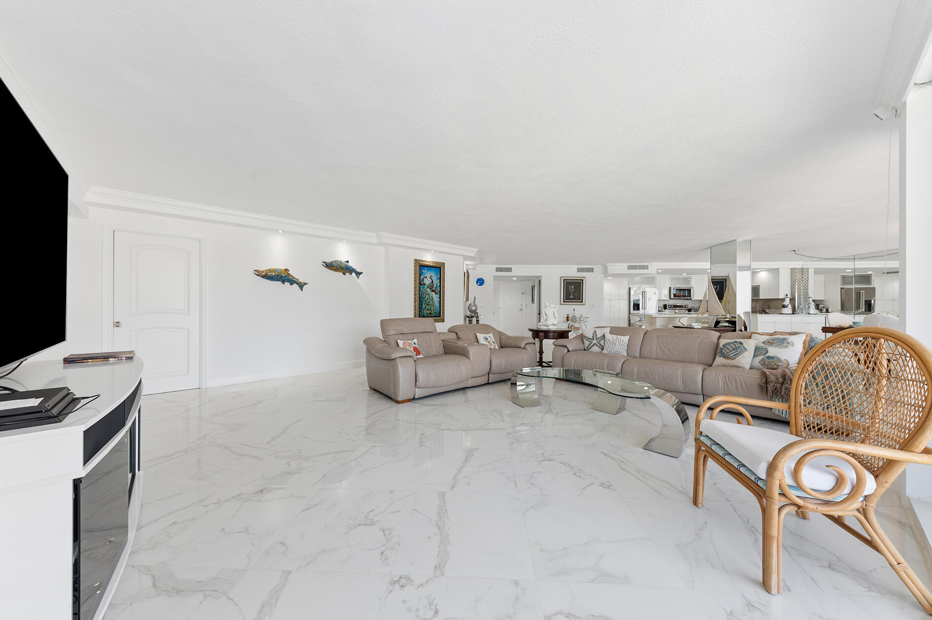 Bright open-plan living room with marble floors, beige sofas, and a white kitchen beyond.