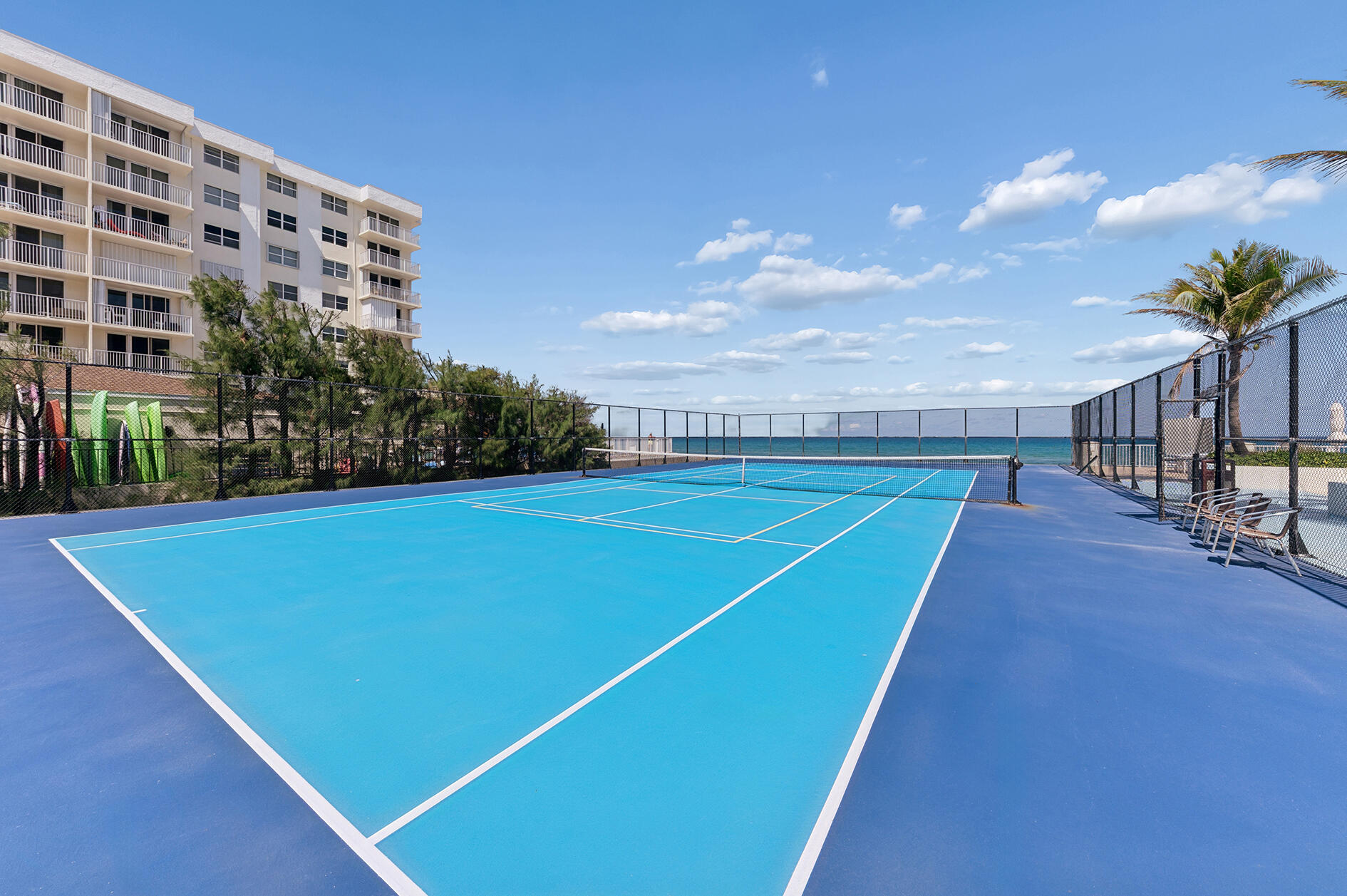 Blue tennis court beside an oceanfront condo, with the Atlantic visible beyond the fence.