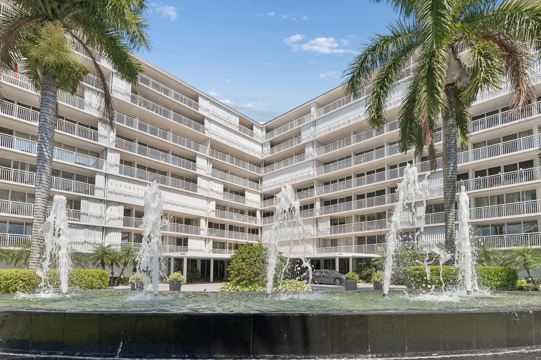 White condominium building with palm trees and a tiered fountain in front.