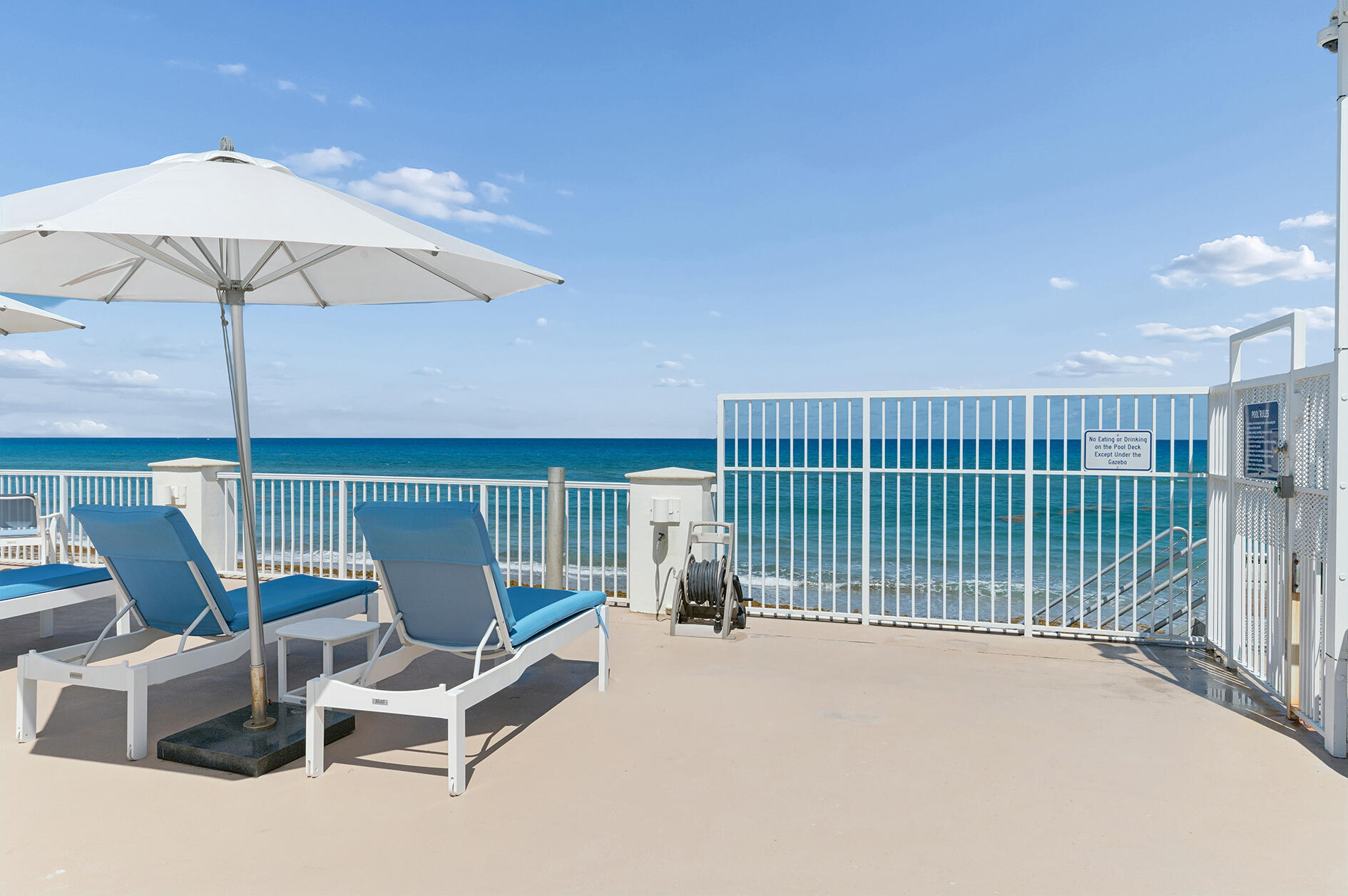 Oceanfront deck with blue lounge chairs, white umbrellas, and a gate opening toward the beach.