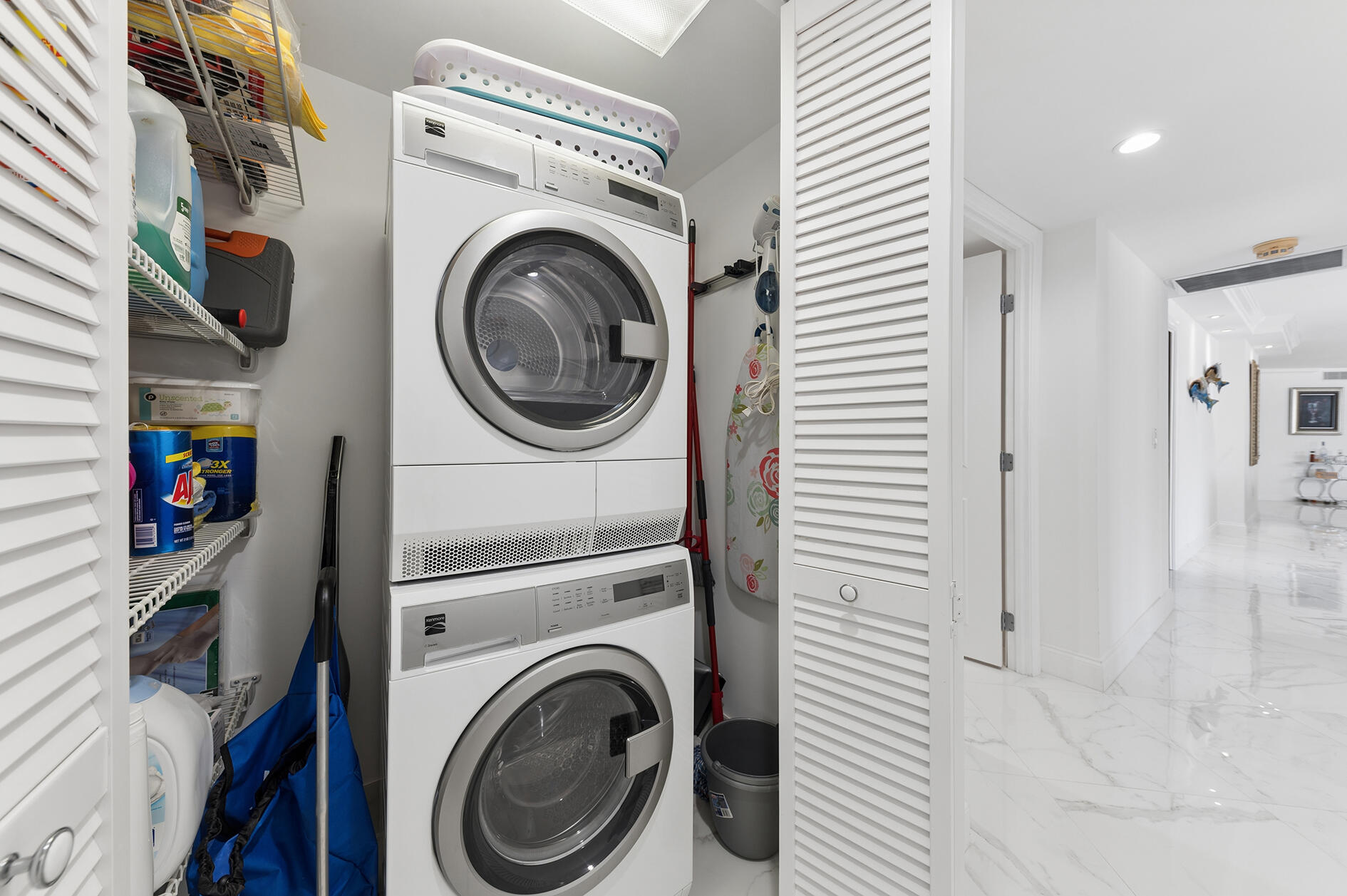 Laundry closet with stacked front-loading washer and dryer, storage shelves, and louvered doors beside a marble-floored hallway.