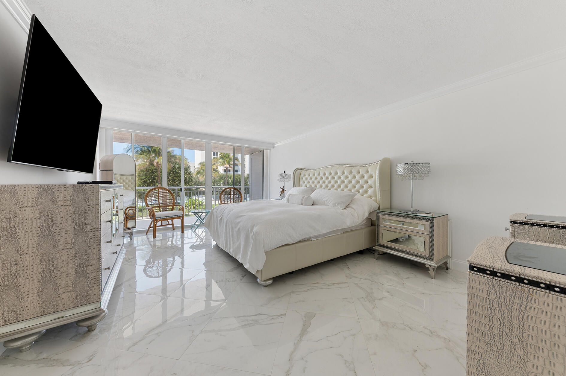 Bright bedroom with a tufted cream bed, marble floors, and sliding doors opening to a palm-lined balcony.
