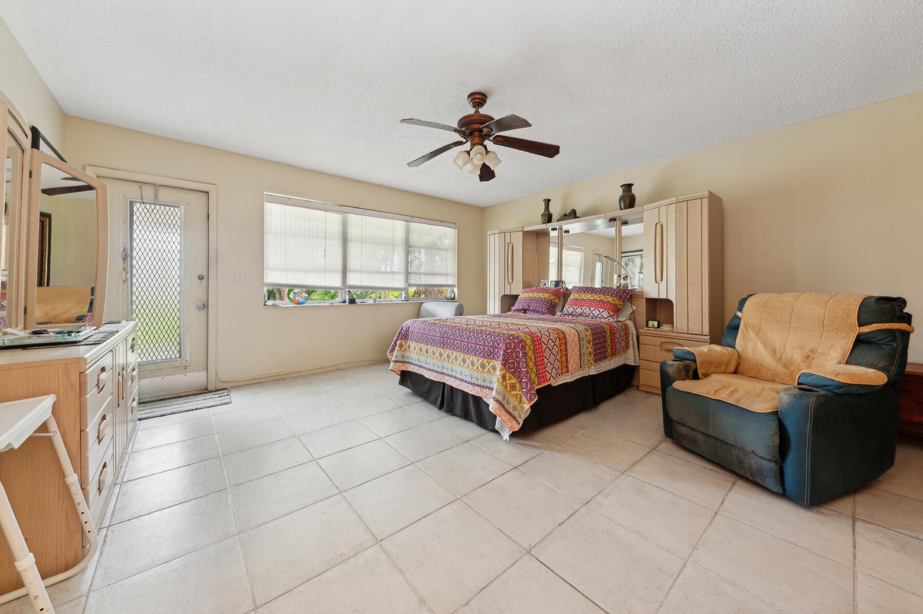 Furnished bedroom with a colorful patterned bedspread, mirrored wardrobe, tiled floor, and cushioned recliner.