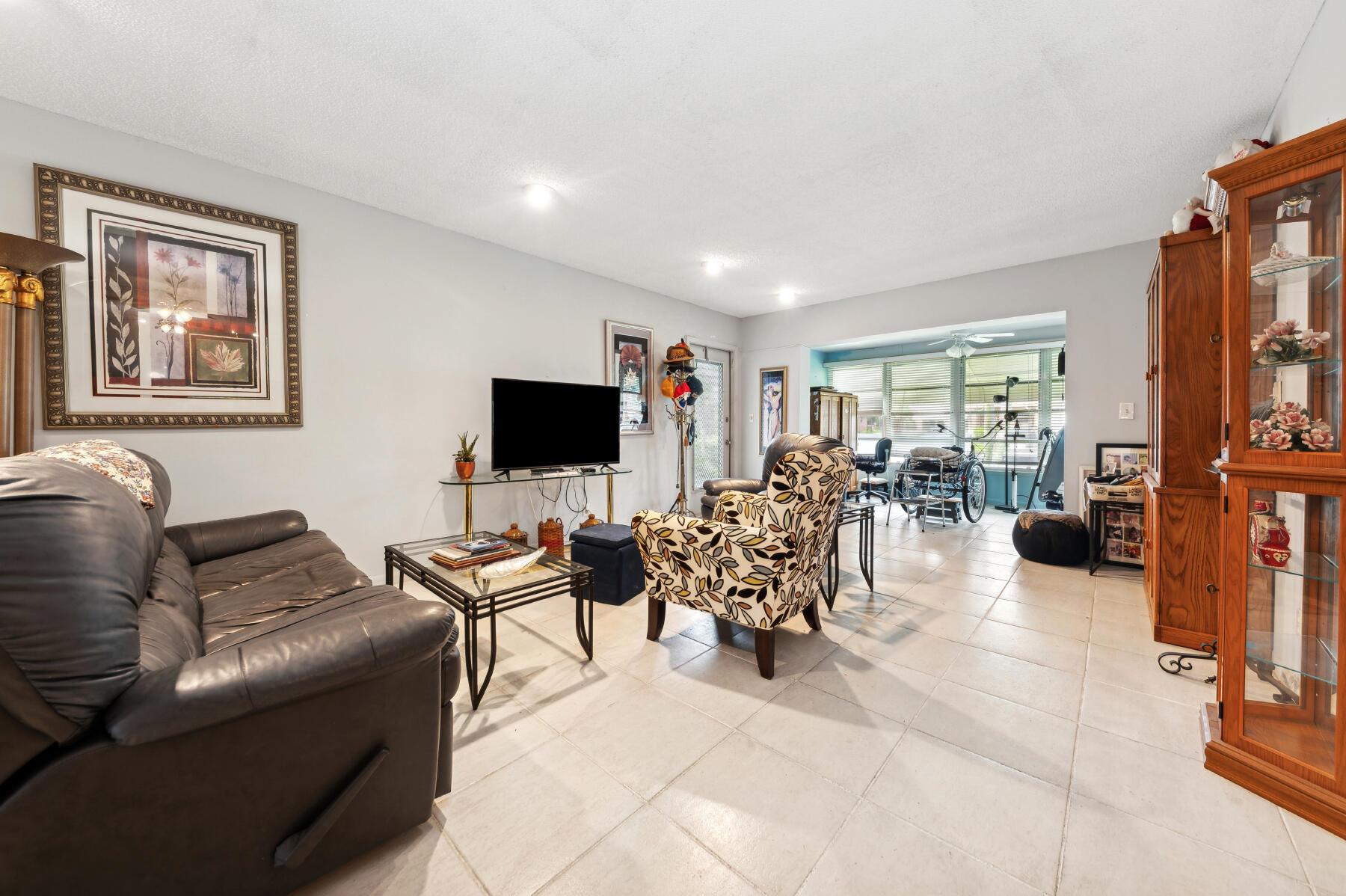 Living room with leather sofa, patterned armchair, tiled floors, and a bright enclosed sunroom beyond.