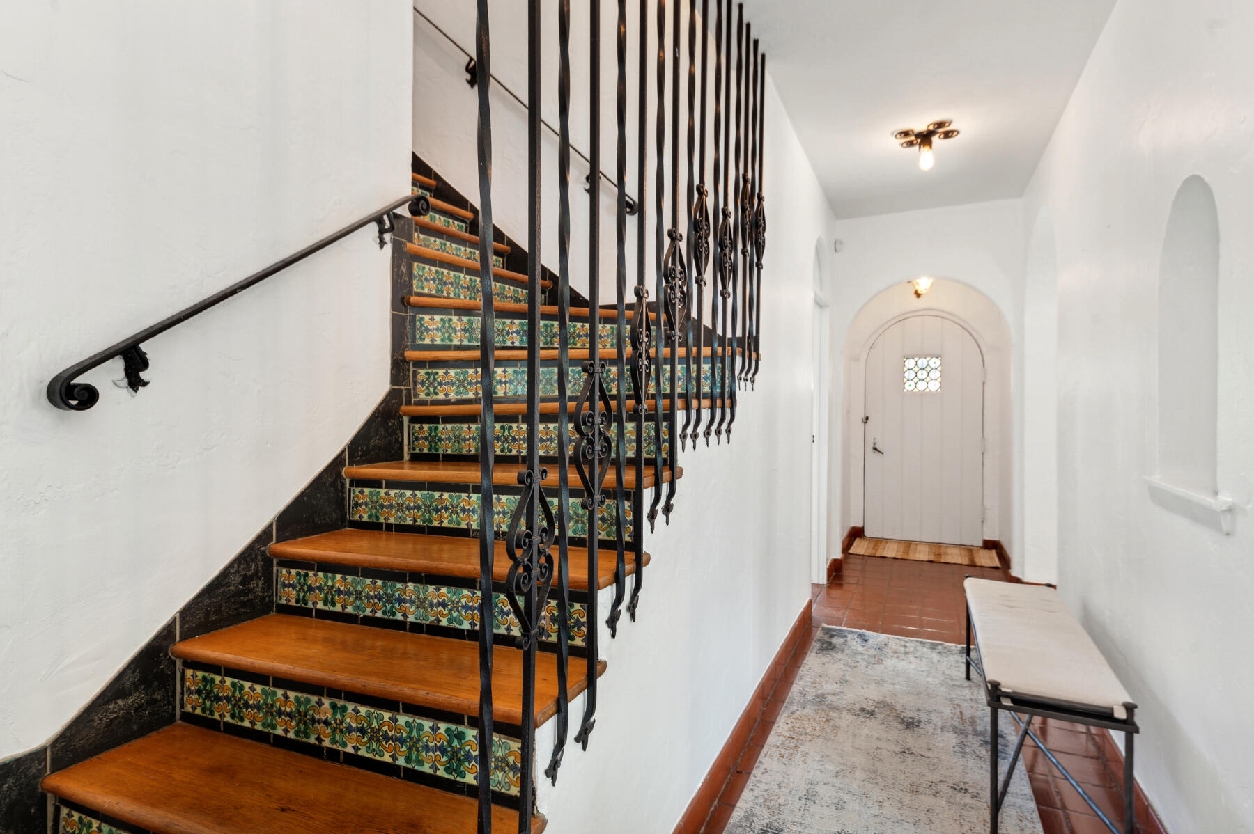 Interior staircase with colorful patterned tile risers and ornate black iron railing beside an arched entryway.