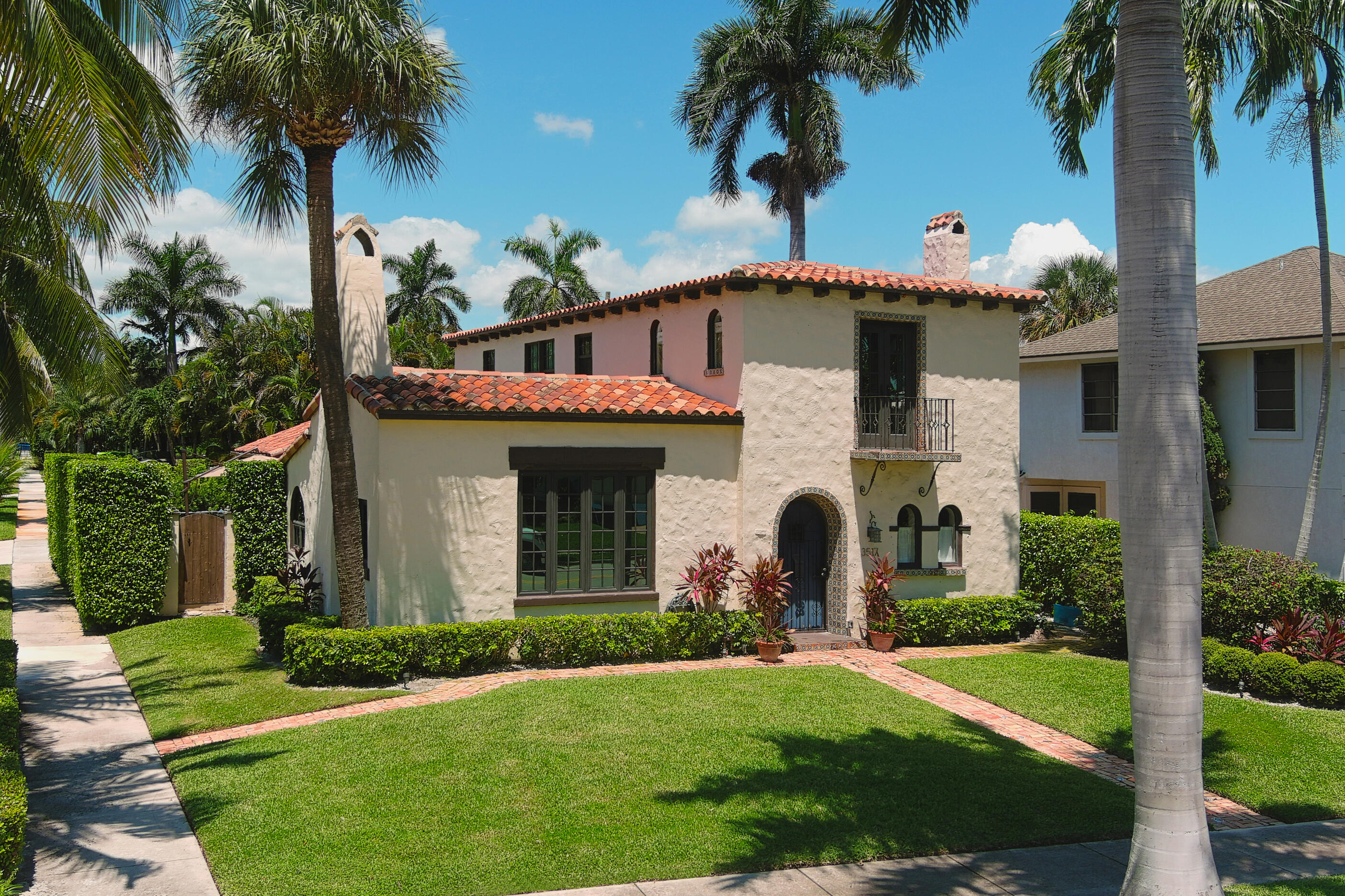 Spanish-style stucco home with terracotta roofs, tiled arched entry, balcony, and palm-framed lawn.