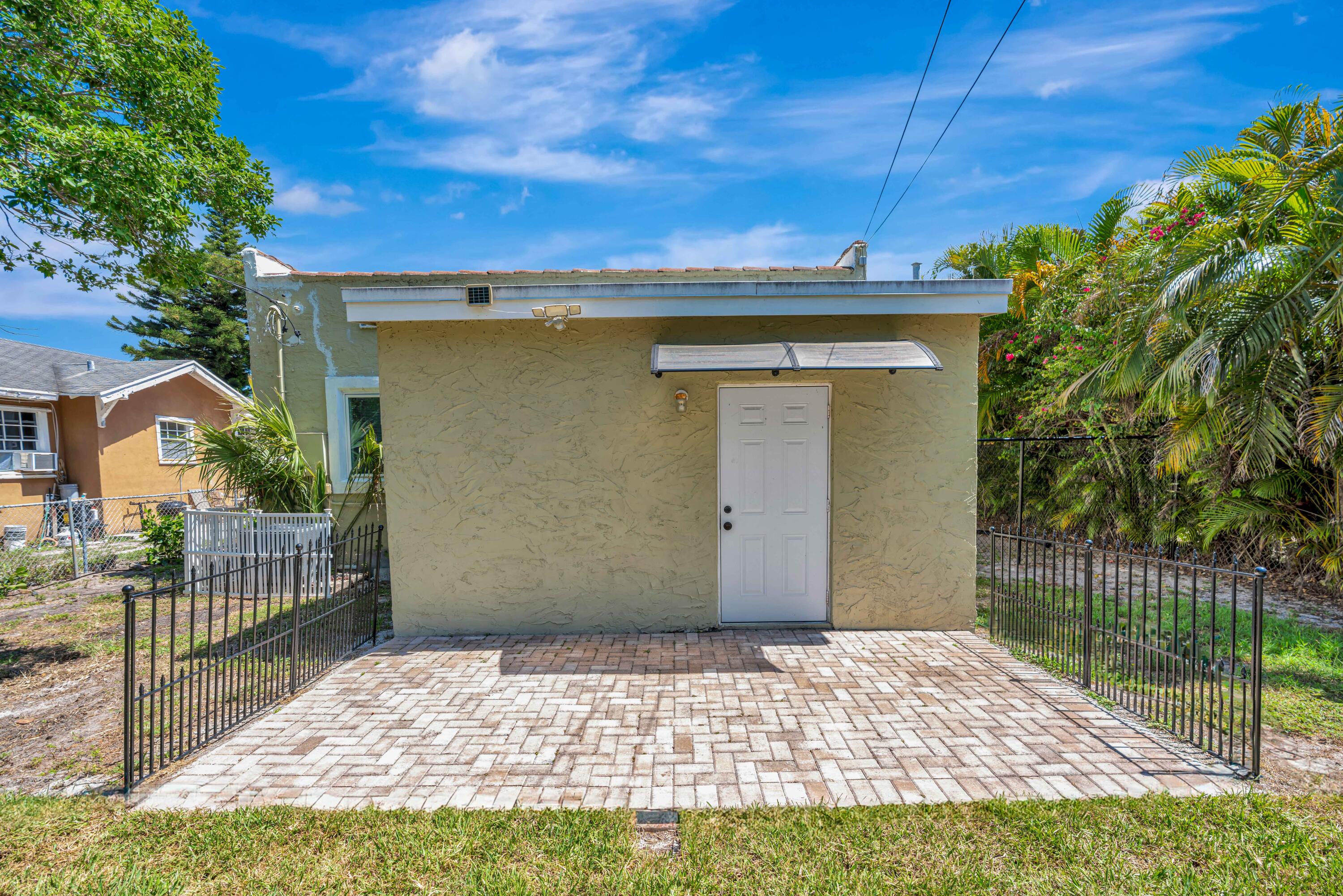 Pale yellow stucco structure with a white door, paved patio, and black metal fencing.