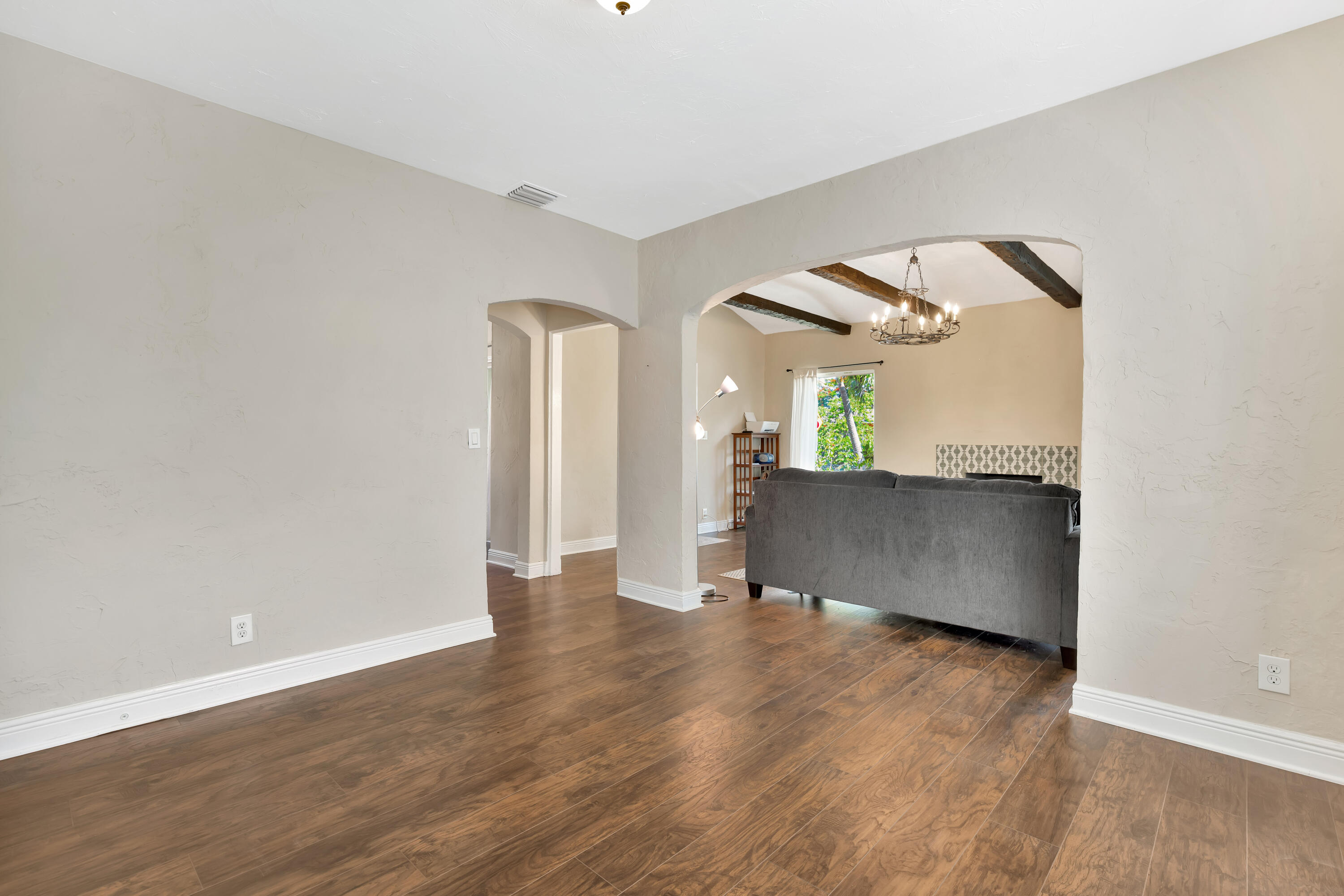 Living room with wood floors, a gray sofa, an arched opening, and exposed ceiling beams.