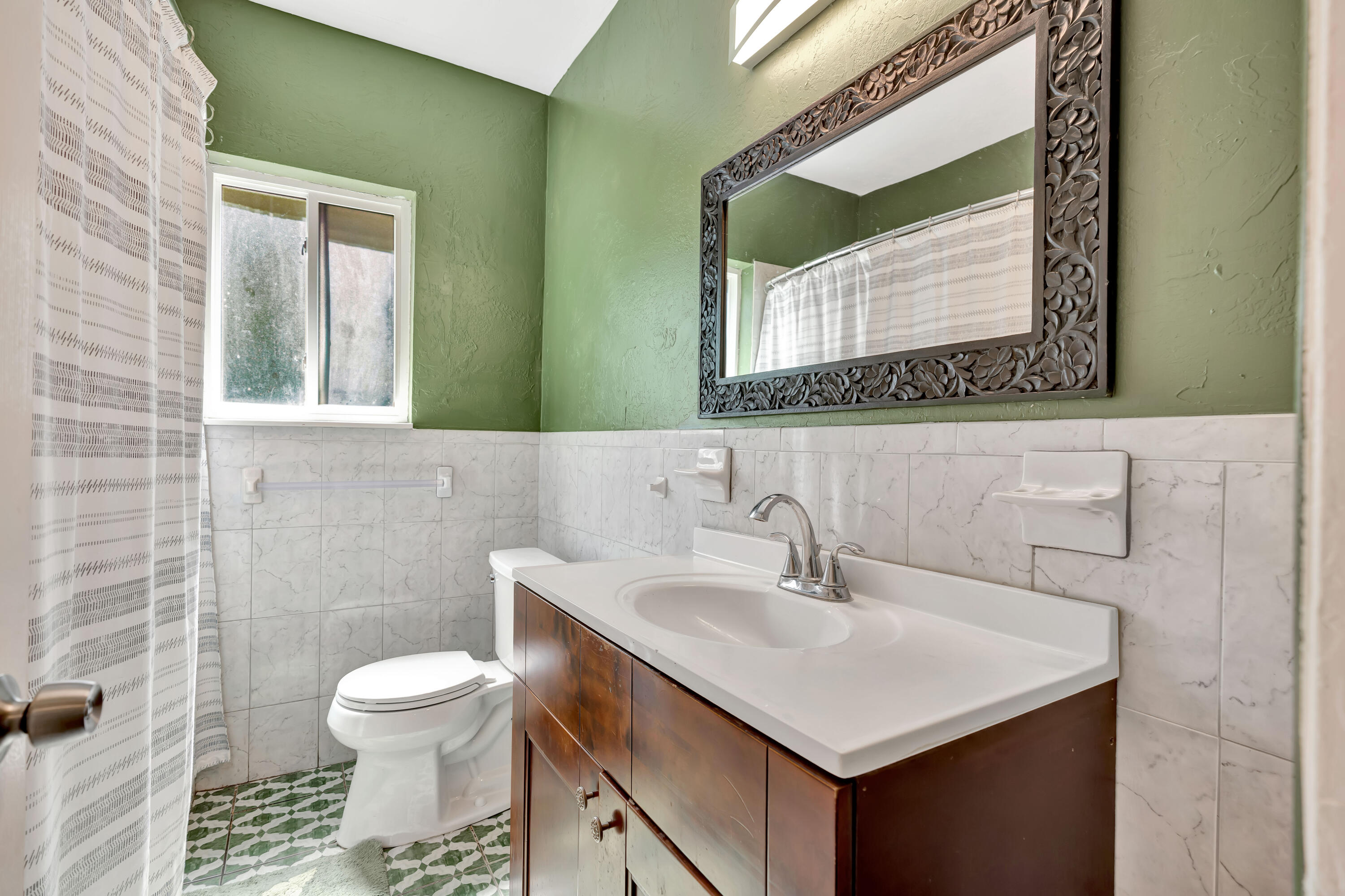 Bathroom with a dark wood vanity, carved mirror, green walls, and patterned tile floor.