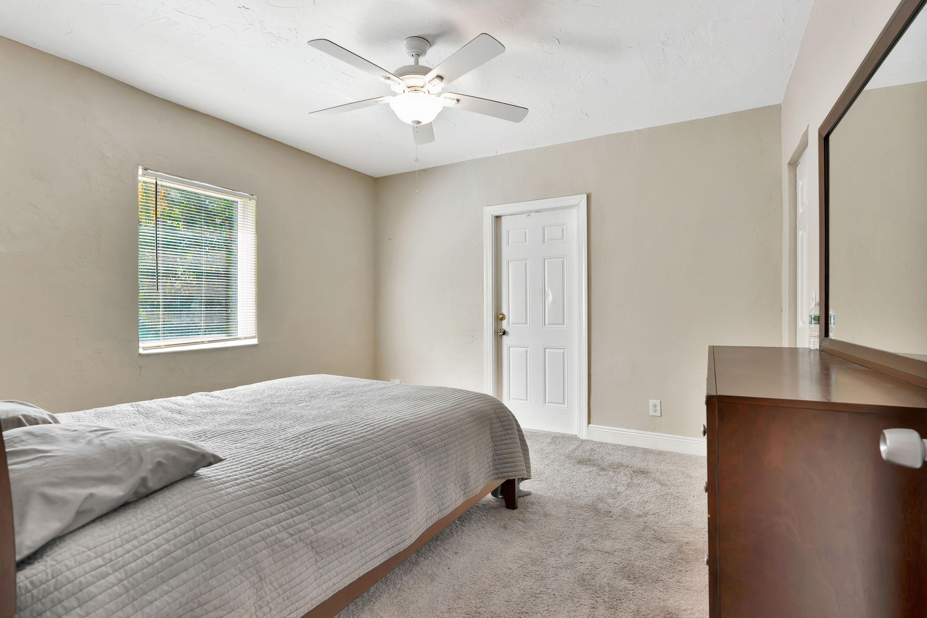 Bedroom with a gray quilted bed, ceiling fan, window blinds, and dark wood dresser.