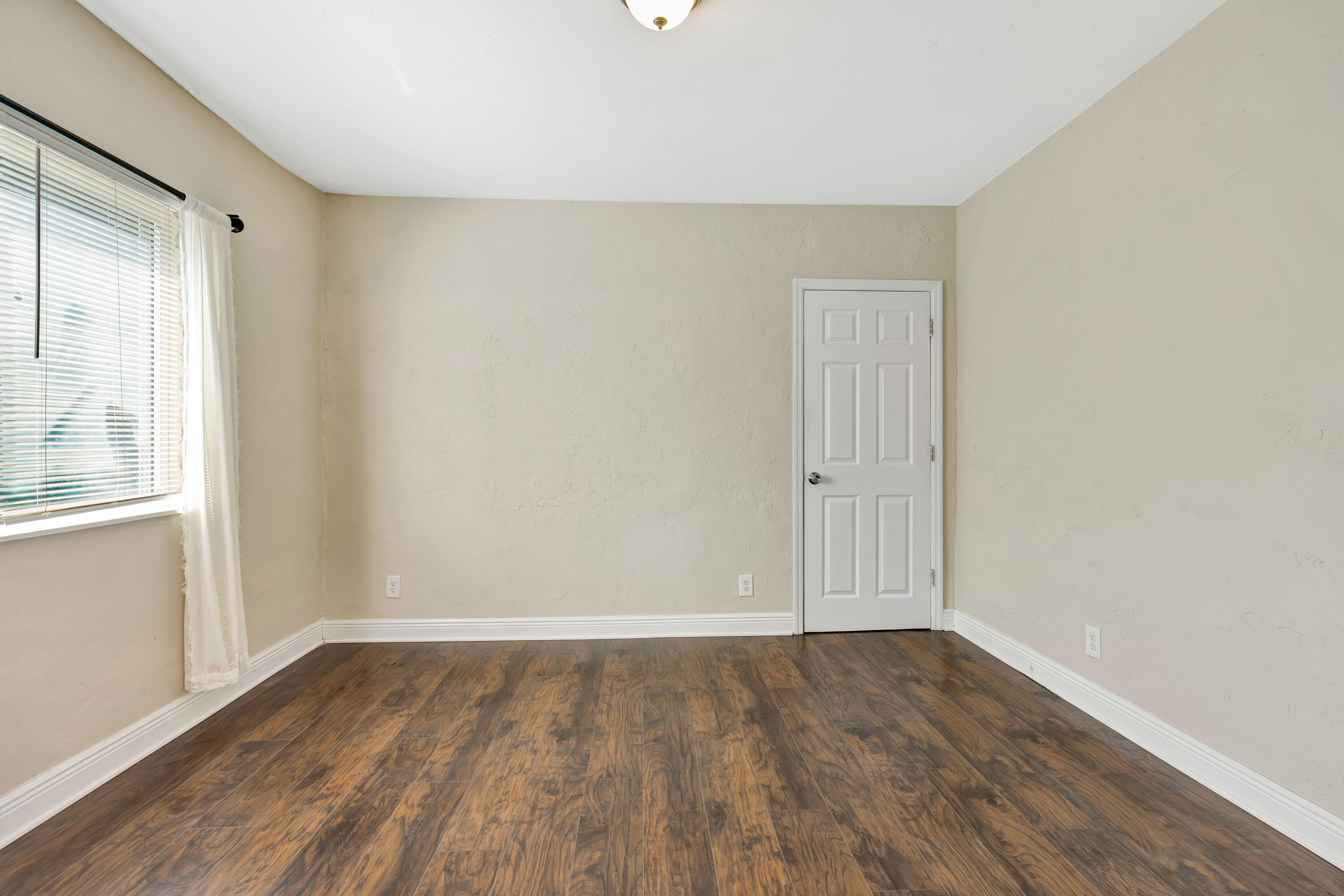 Empty bedroom with dark wood-look flooring, a window with blinds, and a white paneled door.