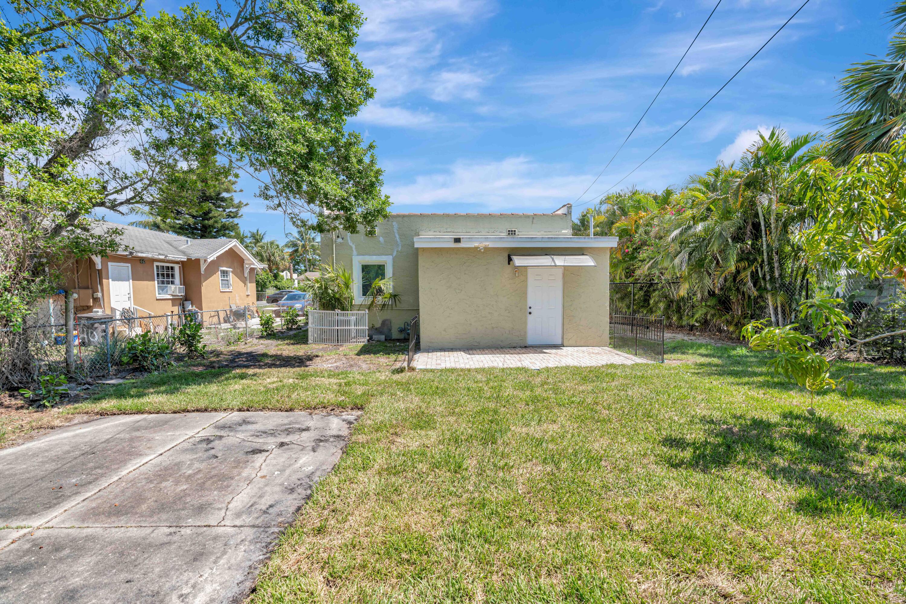 Pale stucco outbuilding with a white door and paved patio beside a grassy, palm-lined yard.