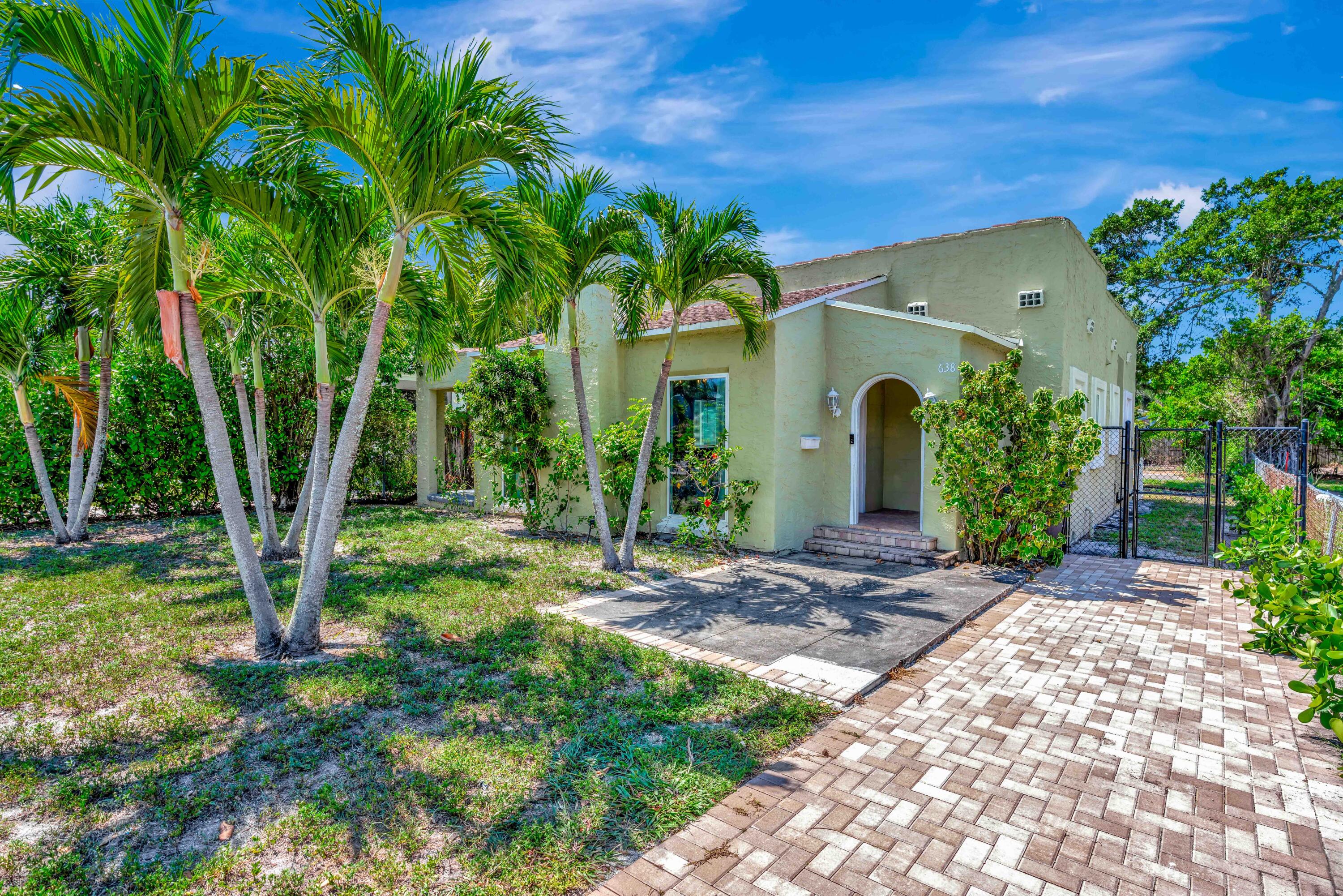Pale green stucco home with an arched entrance, palm trees, and a brick-paver driveway.