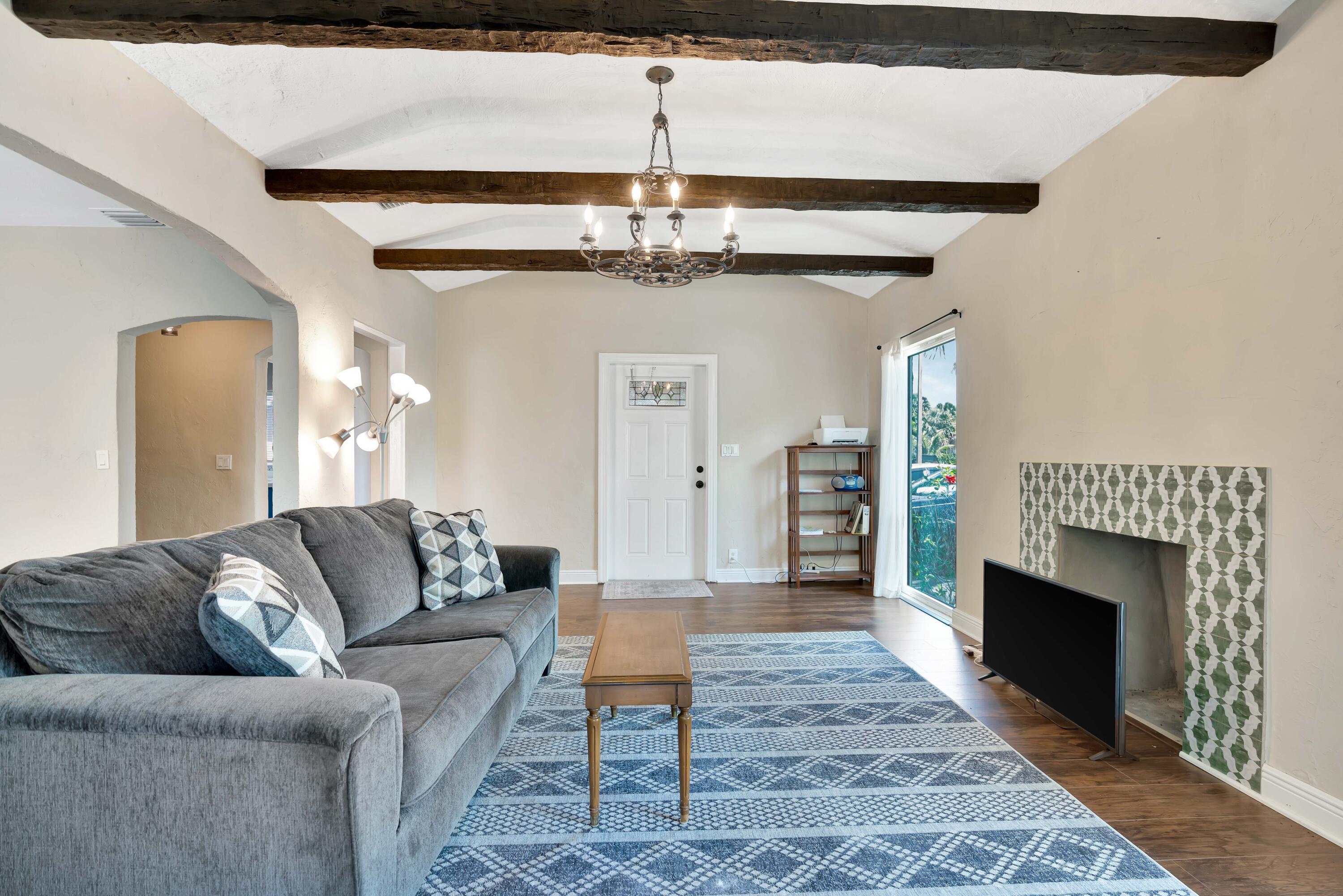 Living room with exposed wood beams, a gray sectional, blue patterned rug, and tiled fireplace.