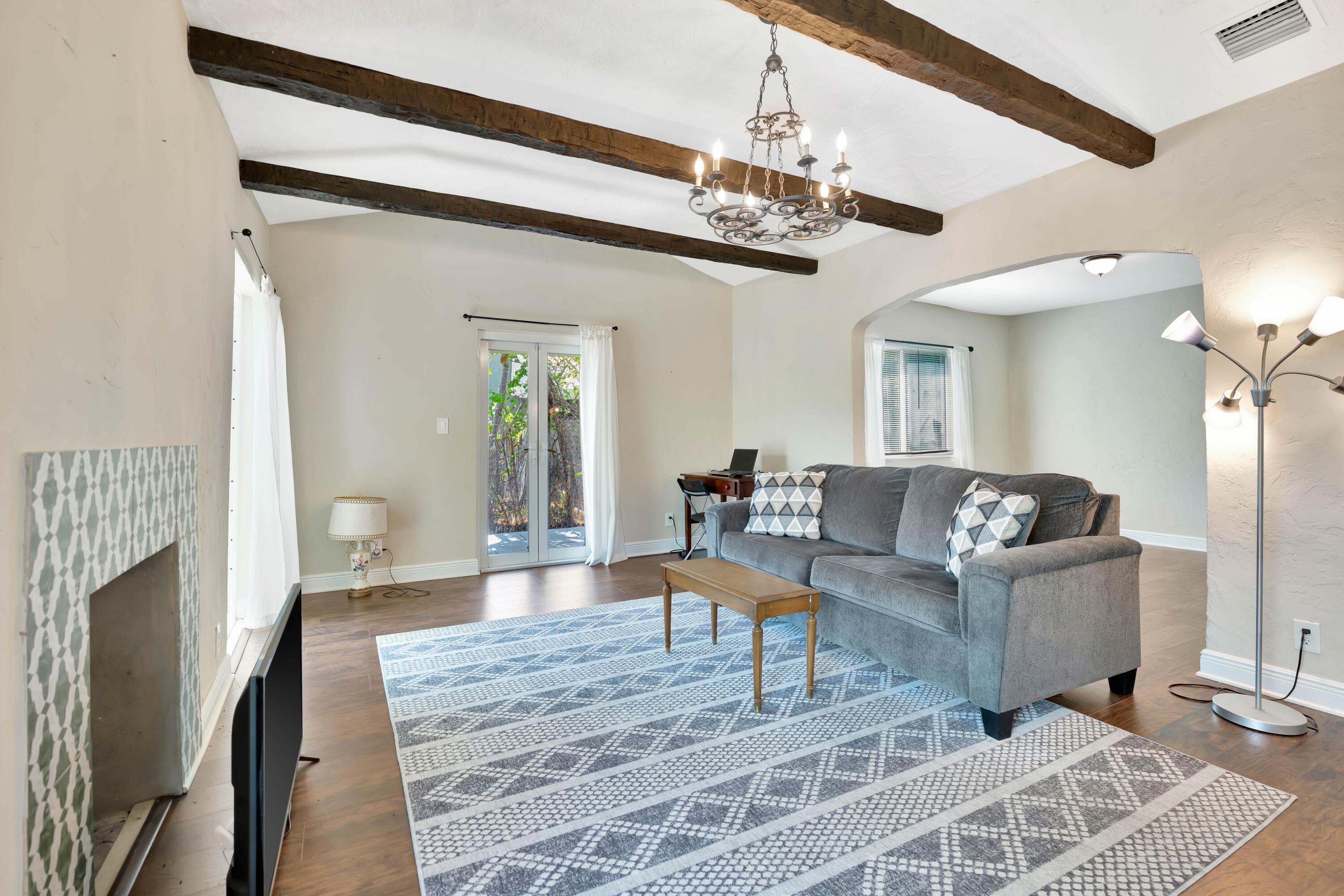 Living room with a gray sofa, patterned rug, tiled fireplace, and exposed dark ceiling beams.