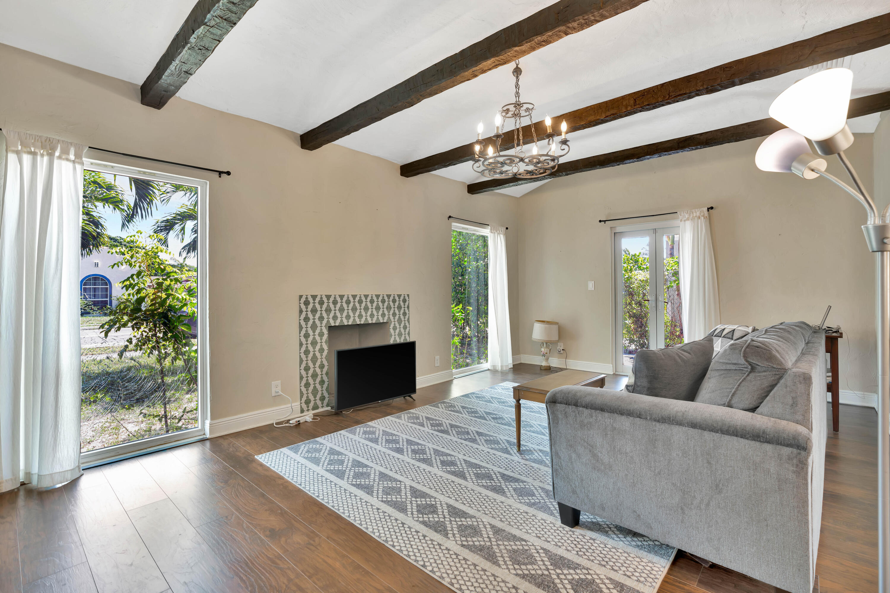 Living room with exposed beams, a patterned-tile fireplace, gray sofa, and large windows overlooking greenery.