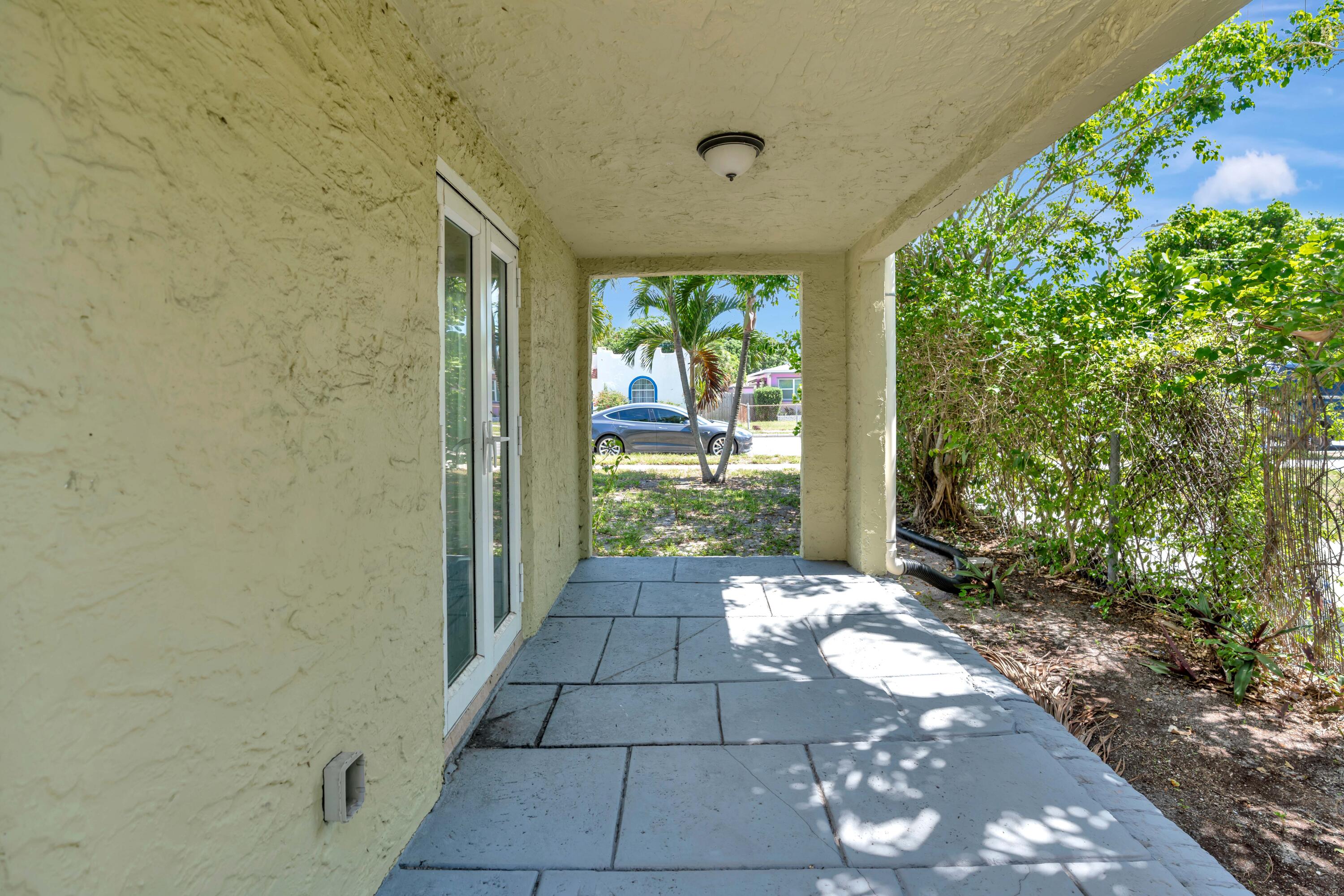 Covered patio with a paved floor, sliding glass doors, and a leafy hedge beside it.