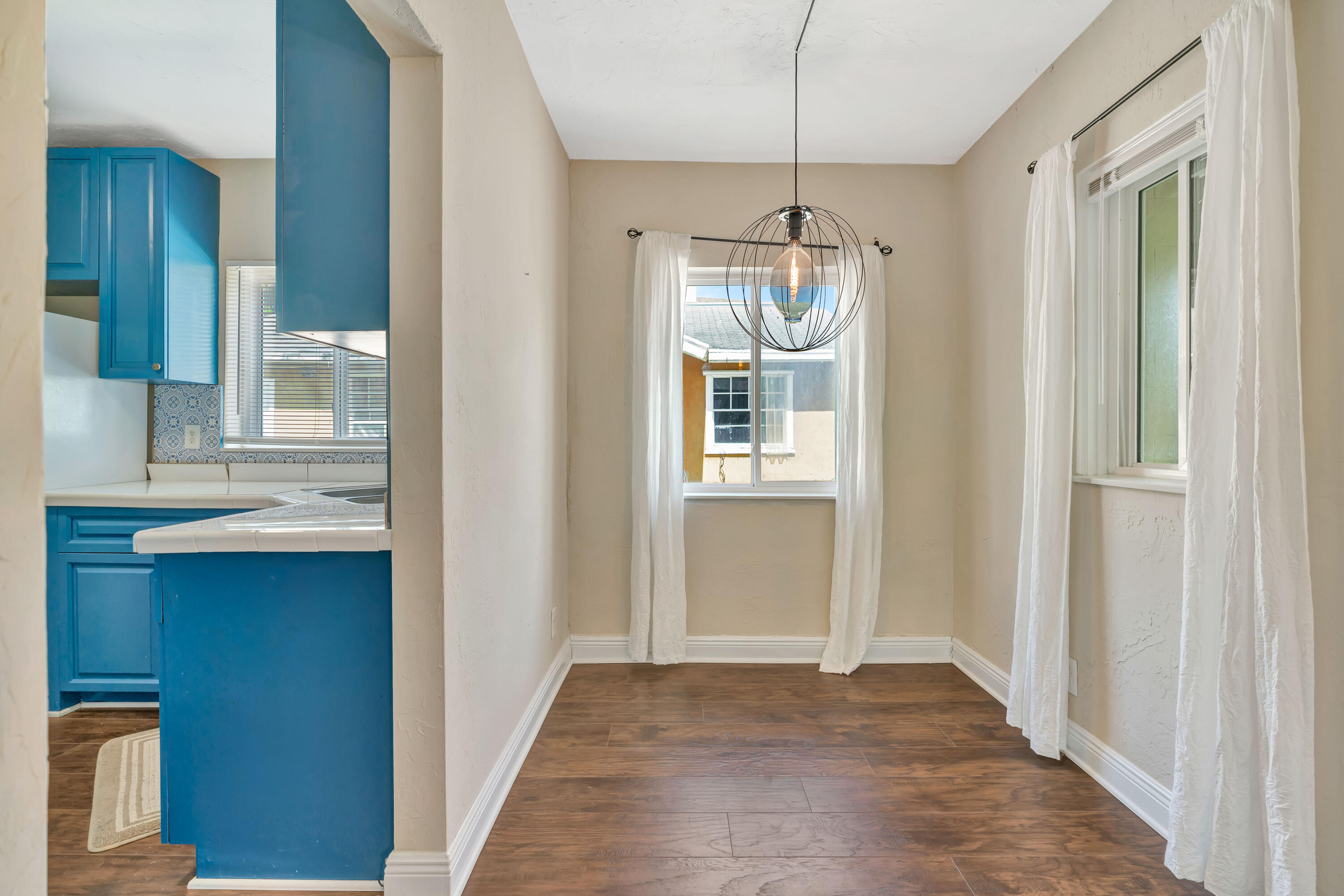 Empty dining area beside a kitchen with blue cabinets and a black wire-cage pendant light.