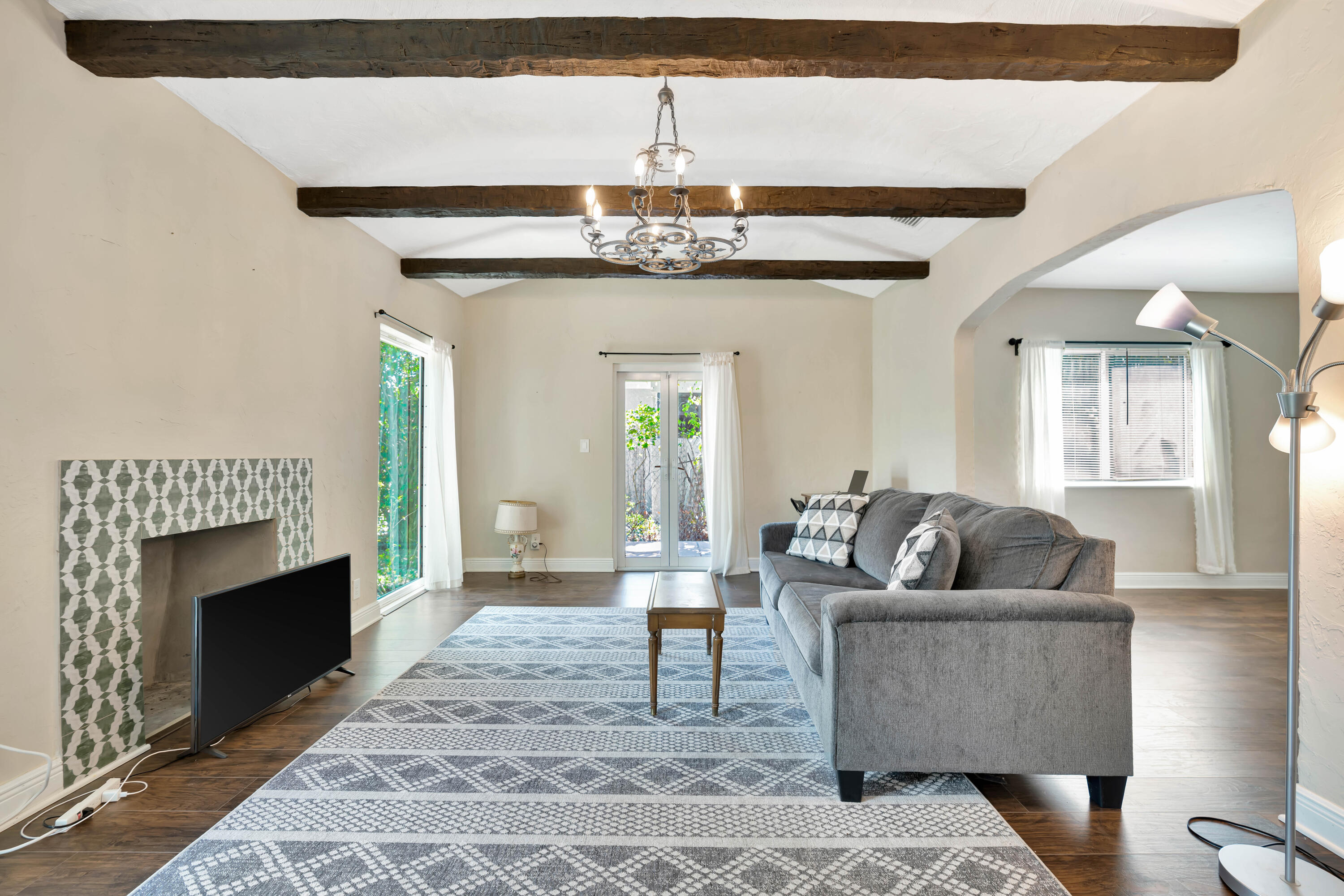 Living room with exposed wood beams, tiled fireplace, gray sofa, and glass doors to greenery.
