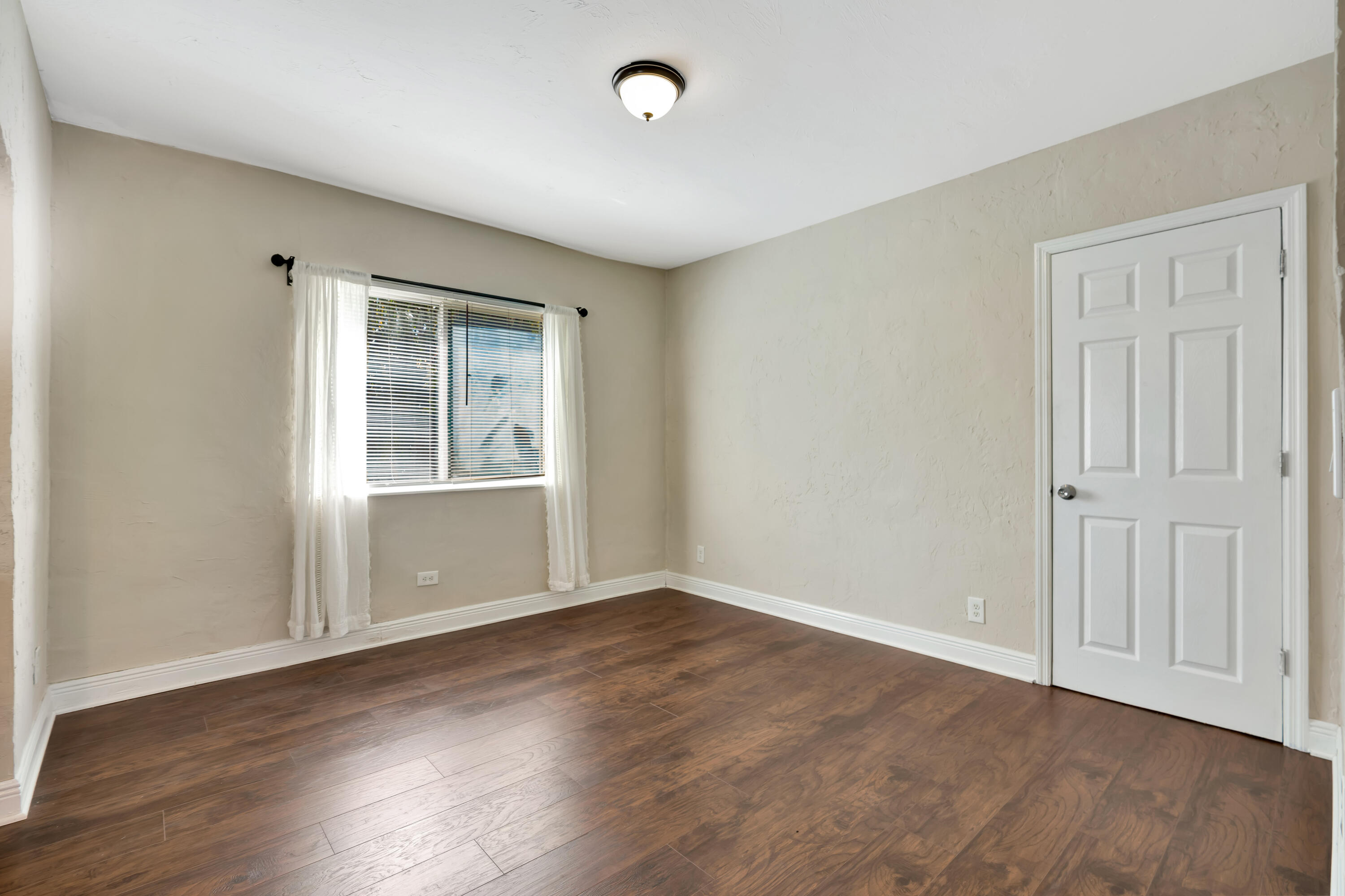 Empty room with dark wood-look floors, a curtained window, and a white paneled door.