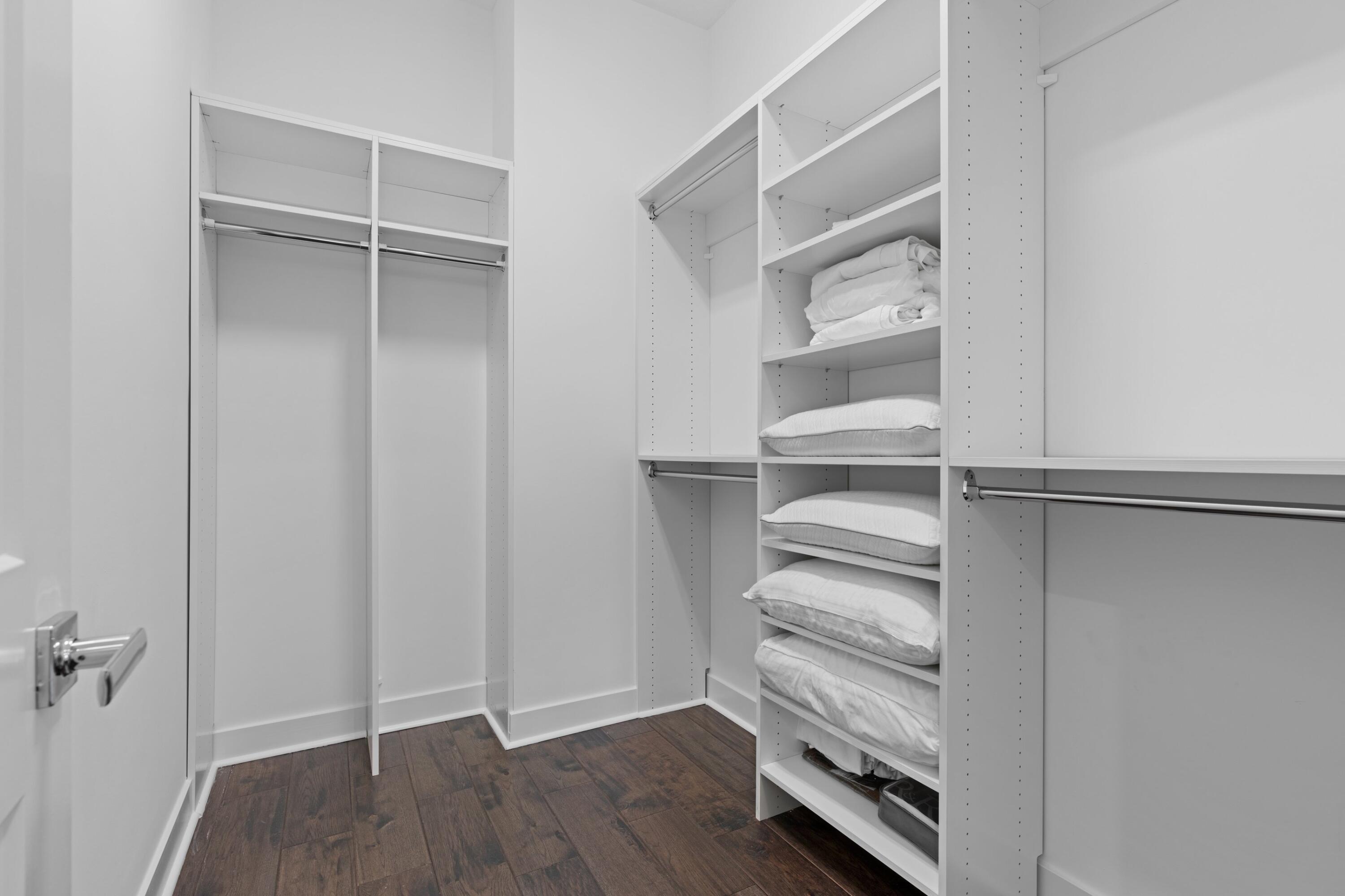 Walk-in closet with white built-in shelves, hanging rods, folded linens, and dark wood-look flooring.