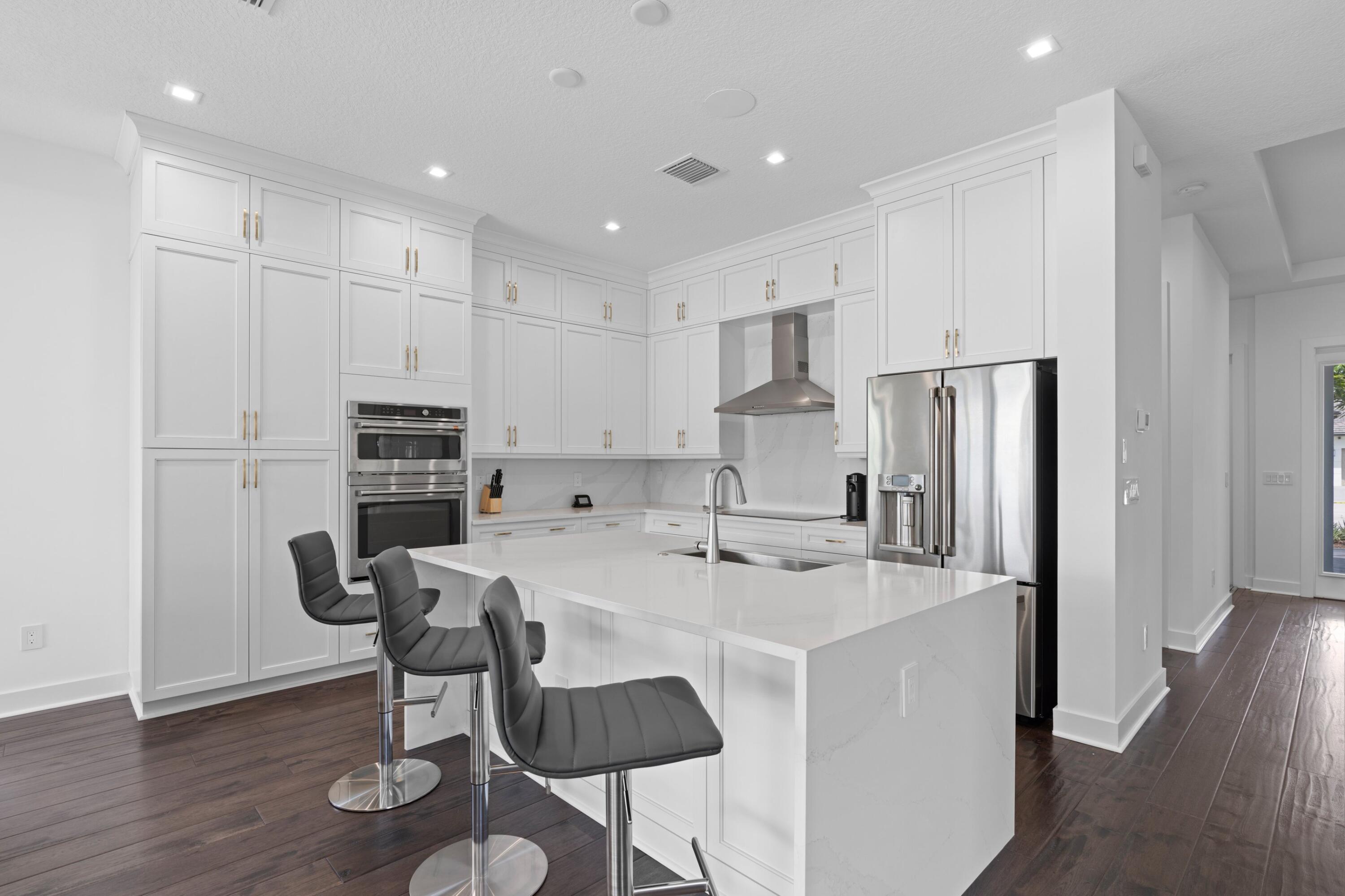 Modern white kitchen with a large island, stainless steel appliances, and gray bar stools.