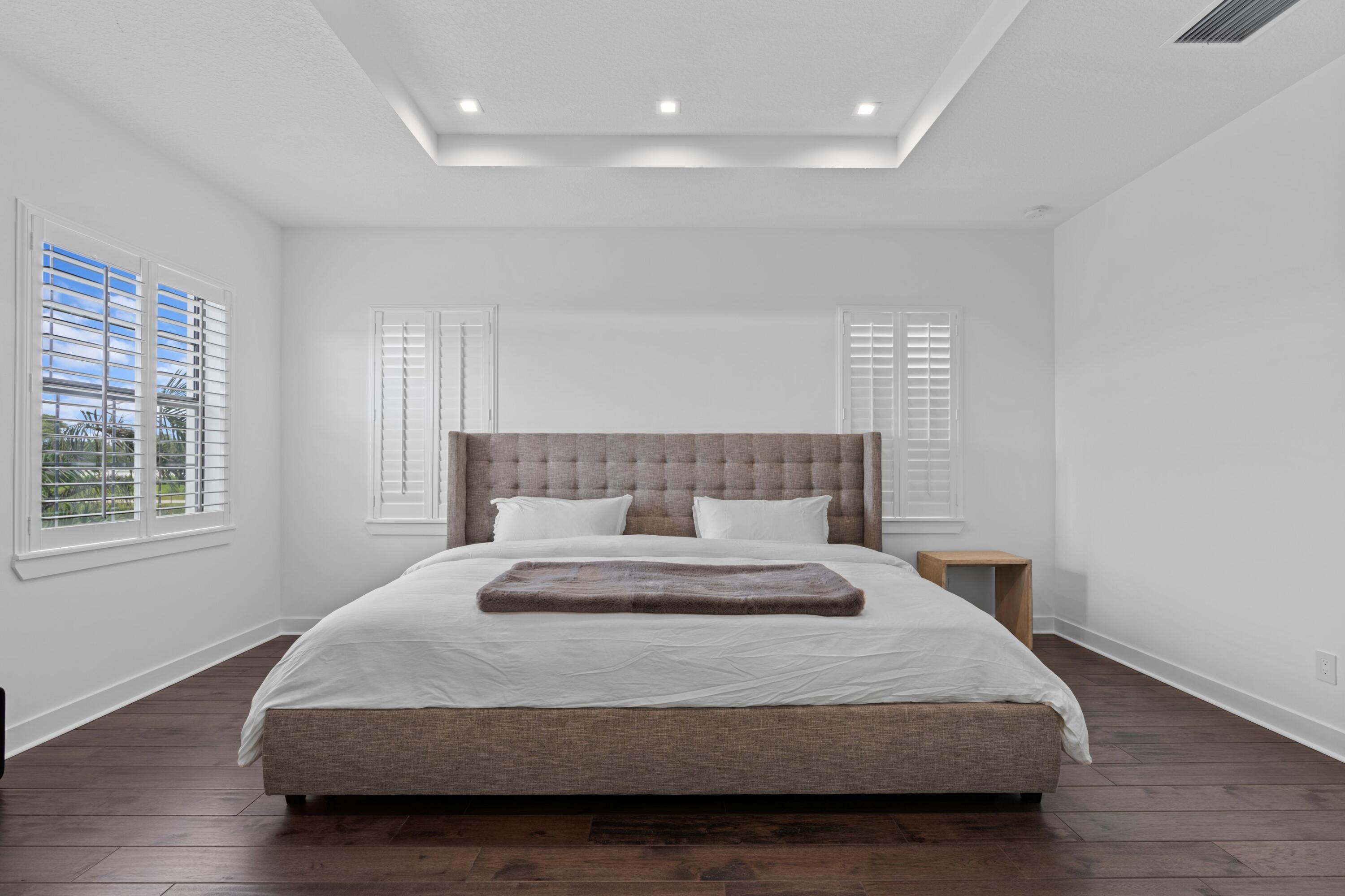 Bedroom with a tufted gray bed, white plantation shutters, dark flooring, and a recessed tray ceiling.