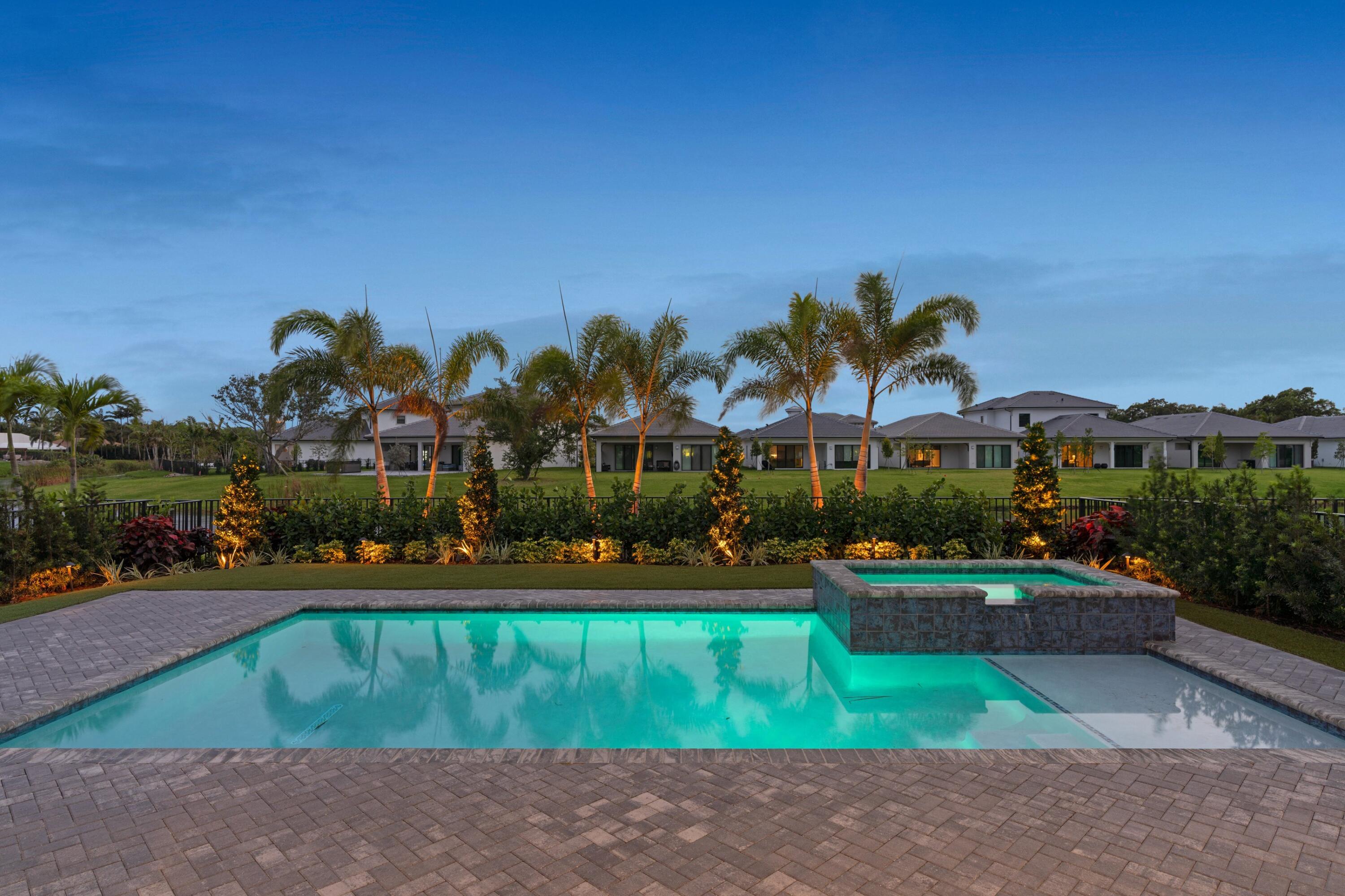 Backyard pool and raised spa with a broad paver deck, illuminated palms, and homes beyond the lawn.