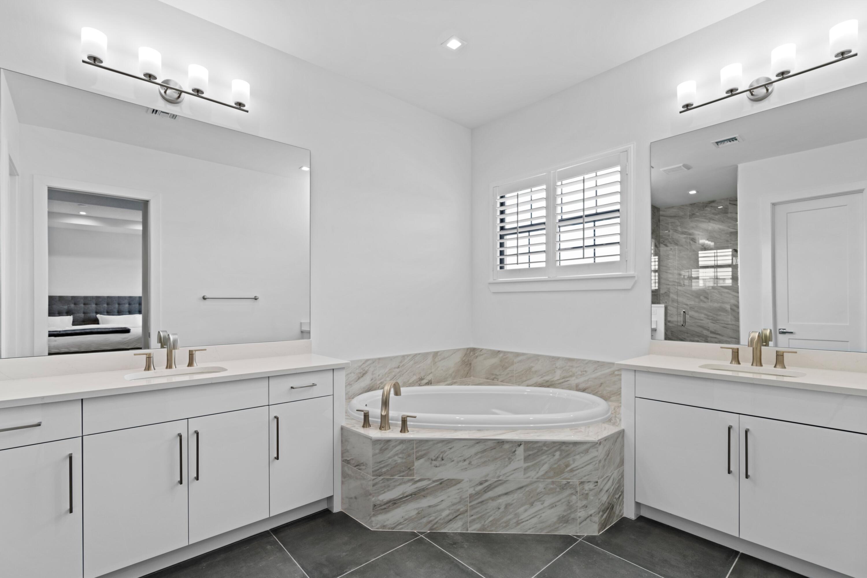 Primary bathroom with a stone-surround soaking tub, twin white vanities, and a shuttered window.
