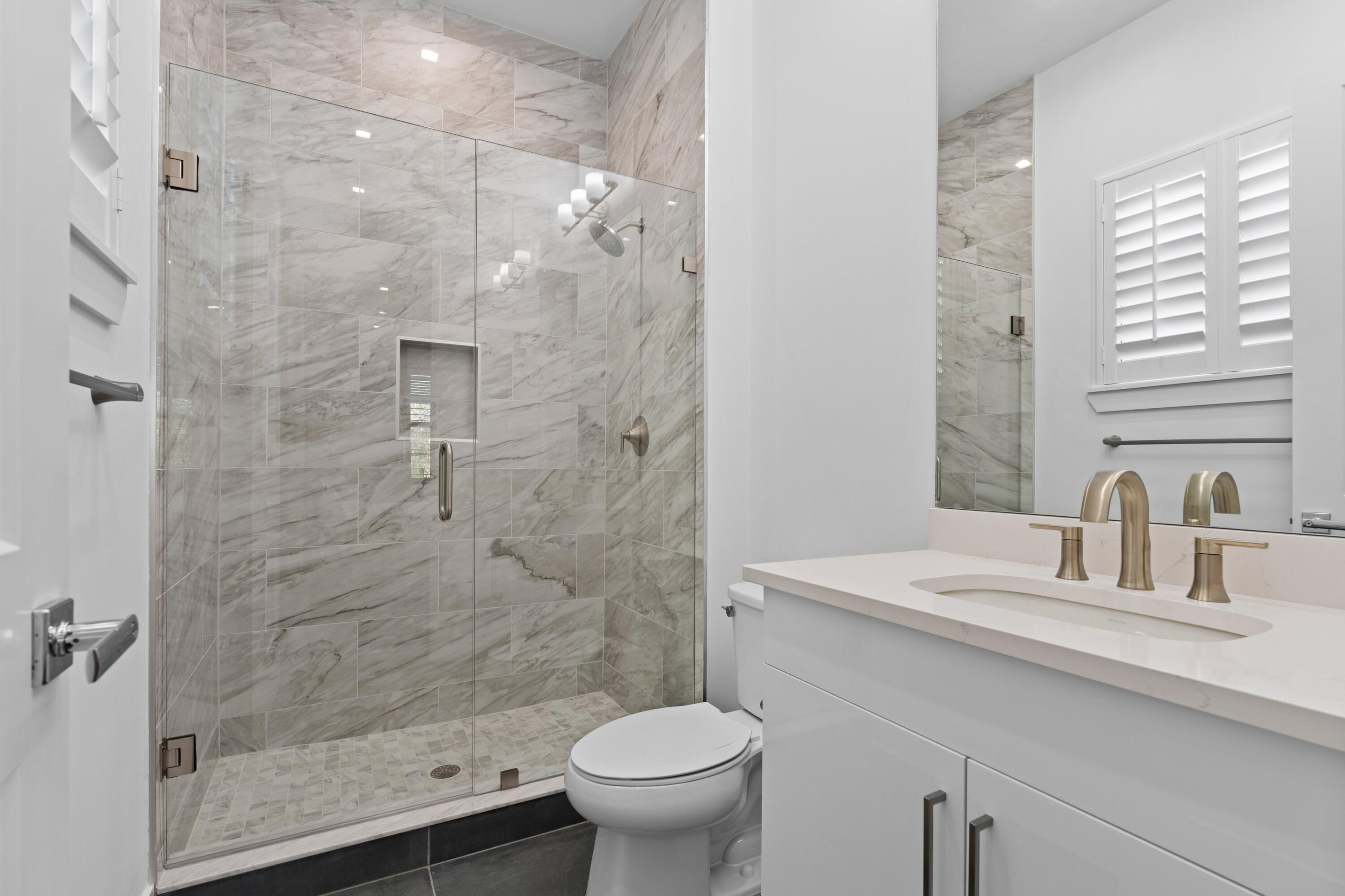 Modern bathroom with a marble-look glass shower, white vanity, brass-tone faucets, and shuttered window.