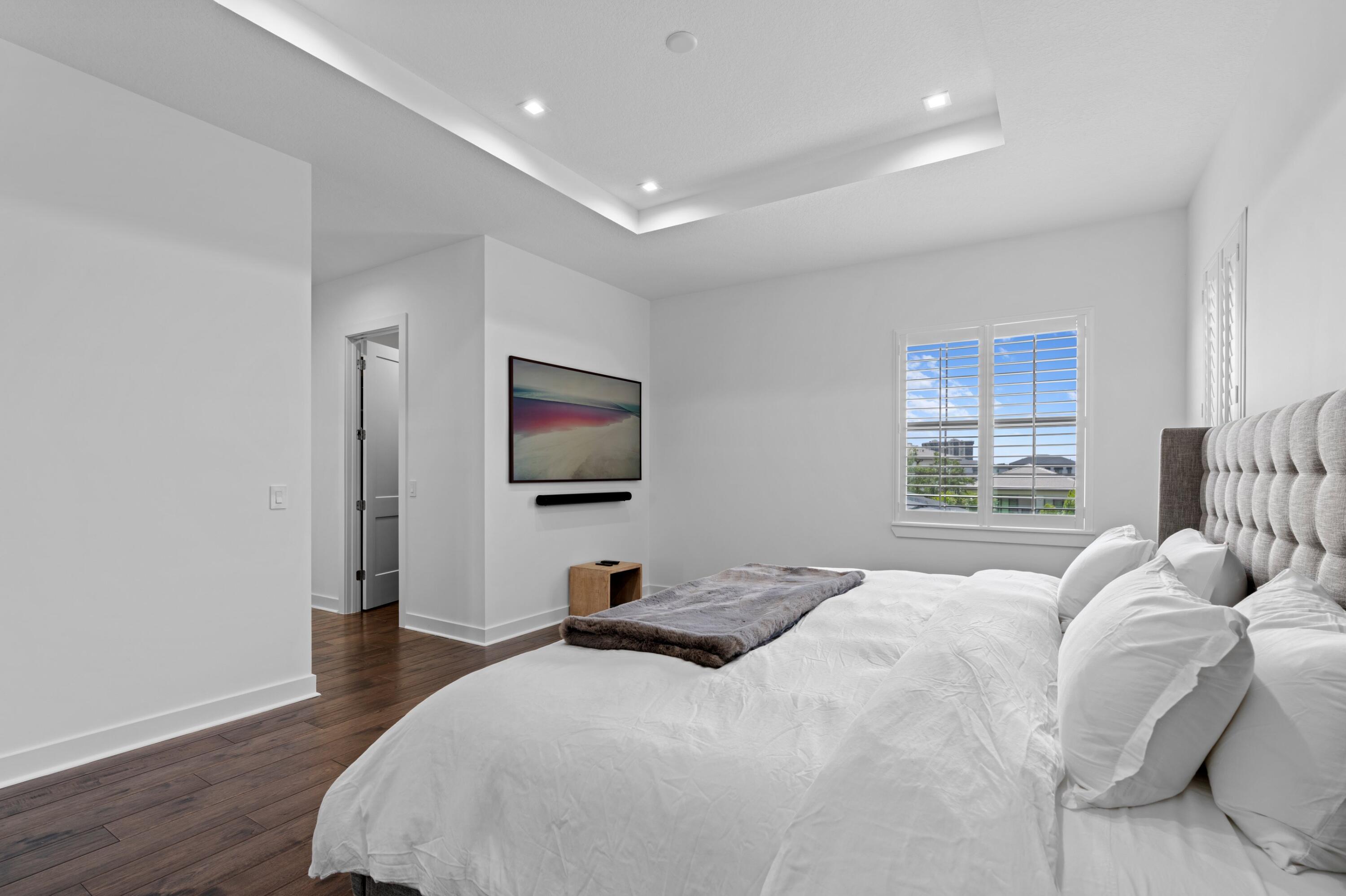 Bedroom with a white bed, tufted gray headboard, dark wood floors, and a shuttered window.