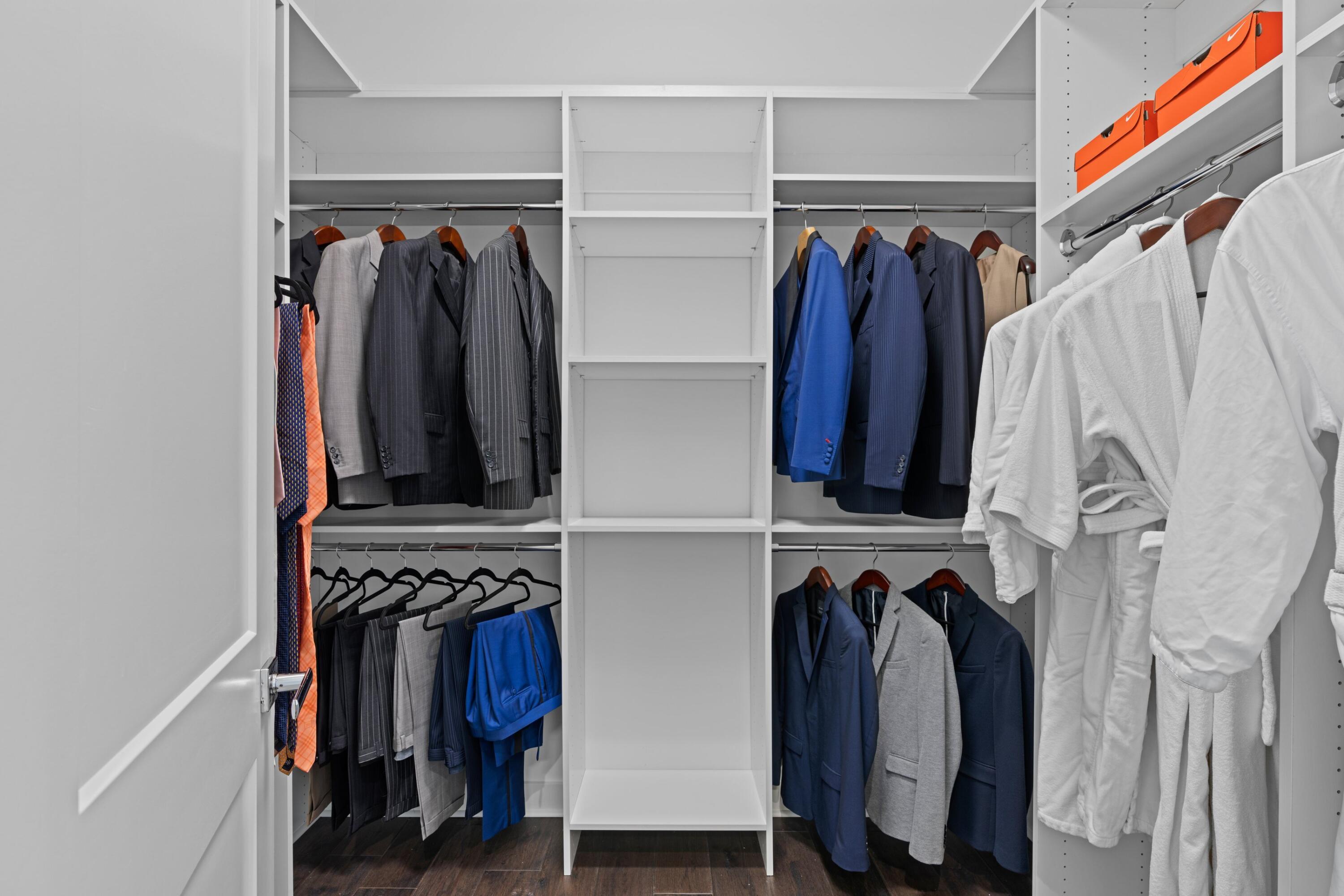 Walk-in closet with white built-in shelving, hanging suits, white robes, and orange storage boxes.