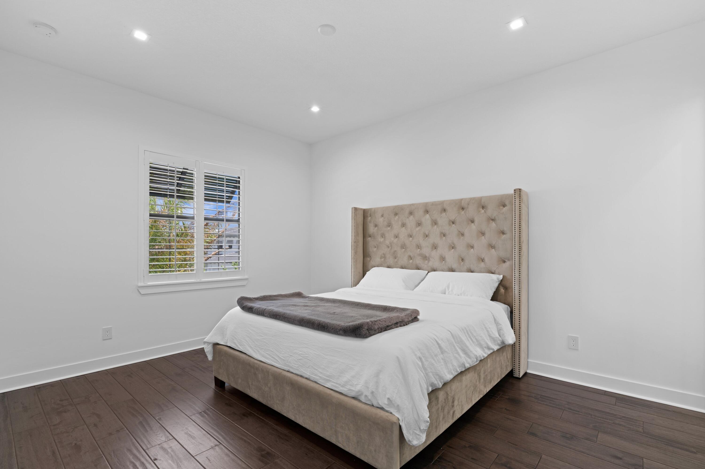 Bedroom with a tall tufted headboard, white bedding, dark wood floors, and a shuttered window.