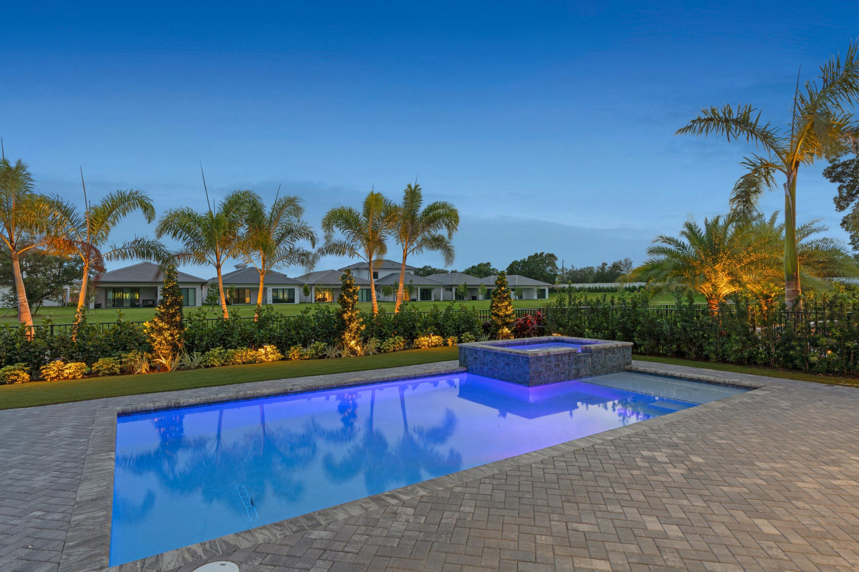 Outdoor pool and raised spa in a paved patio, with illuminated palms and a modern home at dusk.