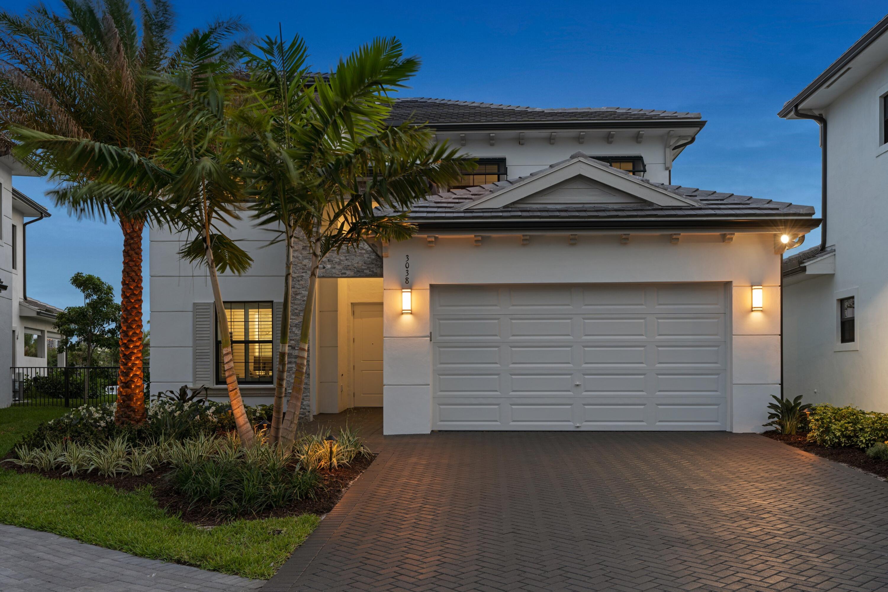 Modern white home with a double garage, palm trees, and a brick-paved driveway at dusk.