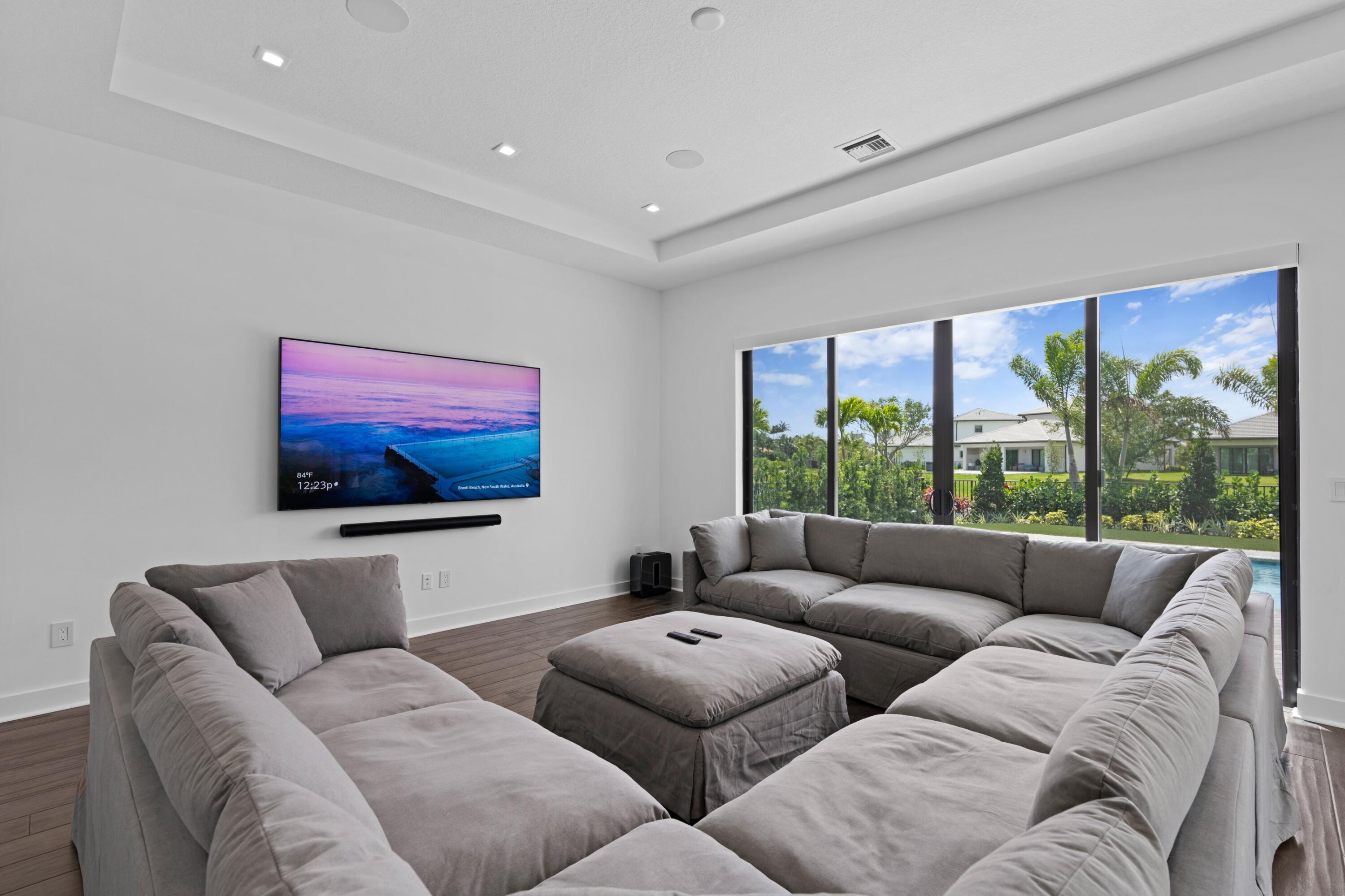 Media room with a large gray sectional, wall-mounted television, and sliding doors overlooking the garden and pool.