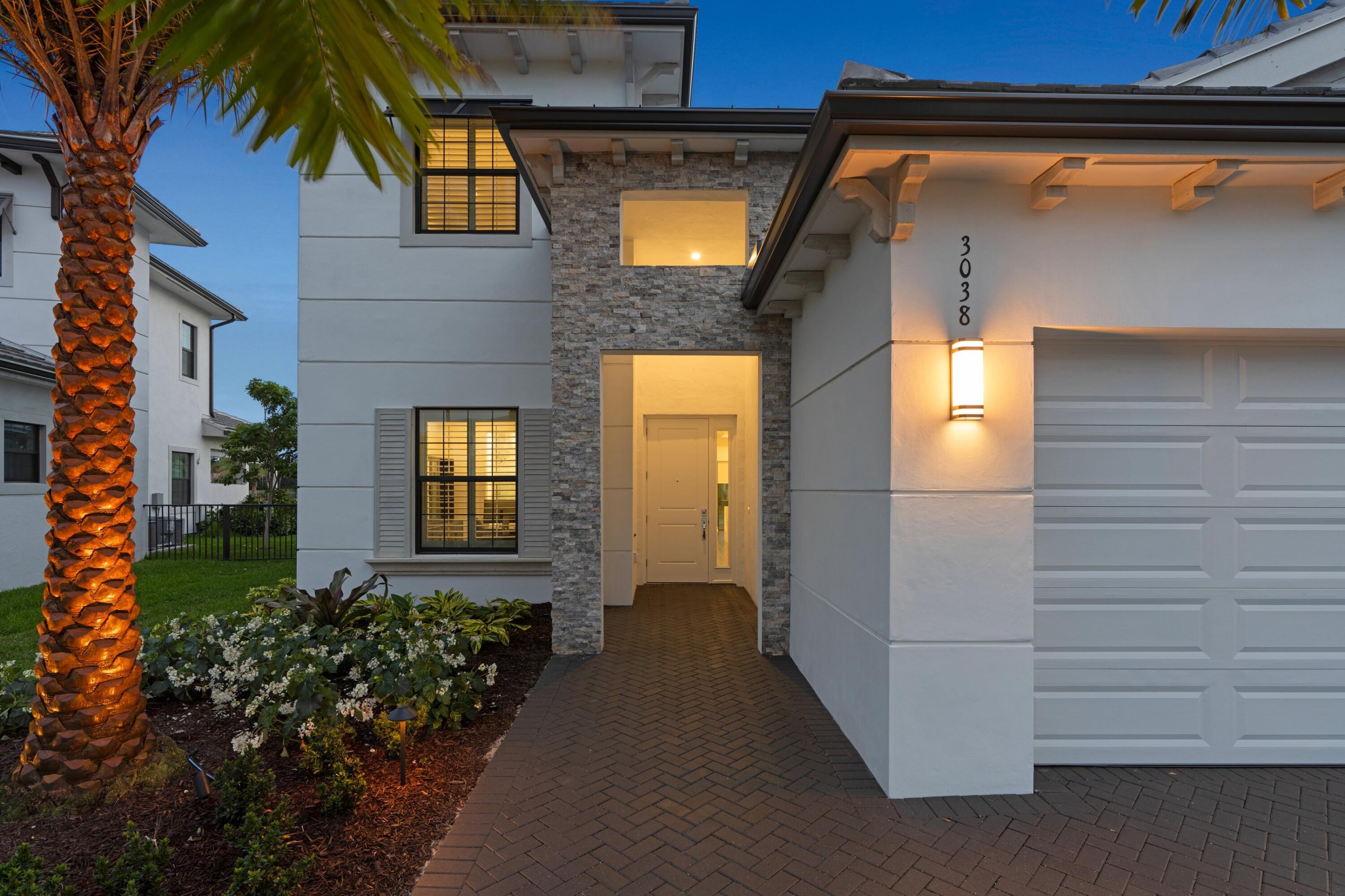 Modern home's stone entry and white garage illuminated at dusk beside a lit palm tree.
