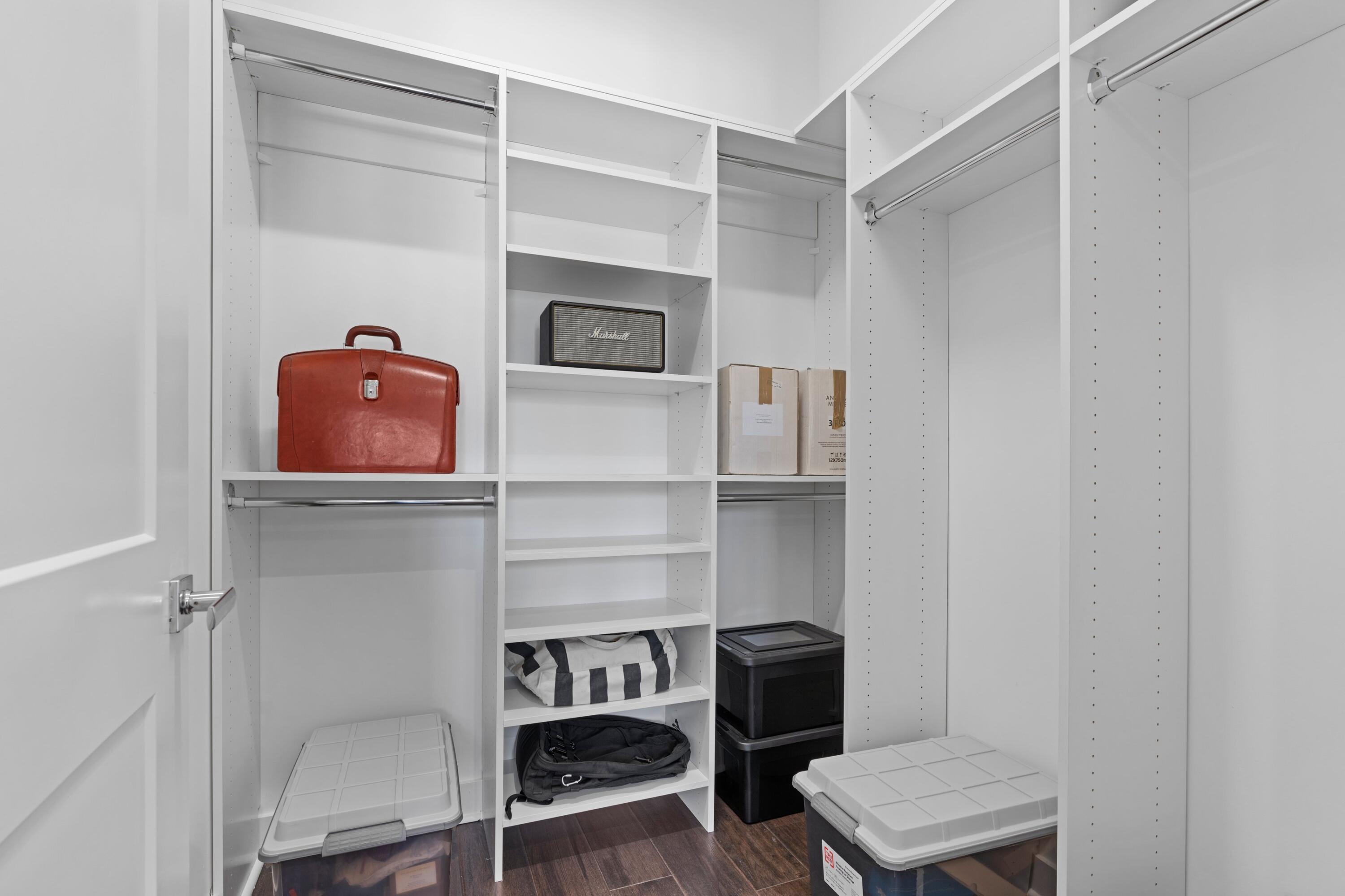 Walk-in closet with white built-in shelving, hanging rods, storage bins, and a brown leather case.
