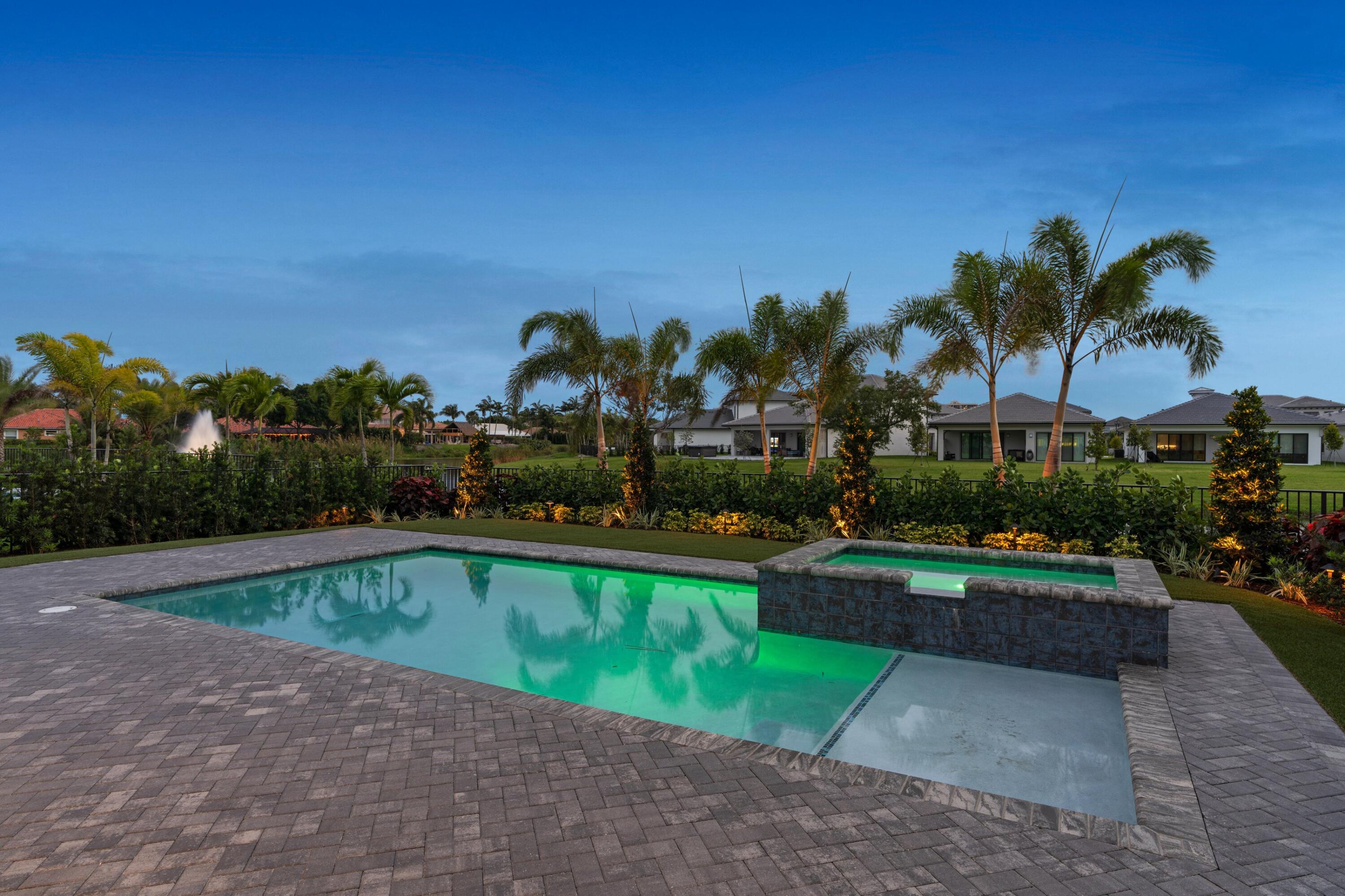 Backyard pool and raised spa with a gray paver deck, tropical palms, and garden lighting at dusk.
