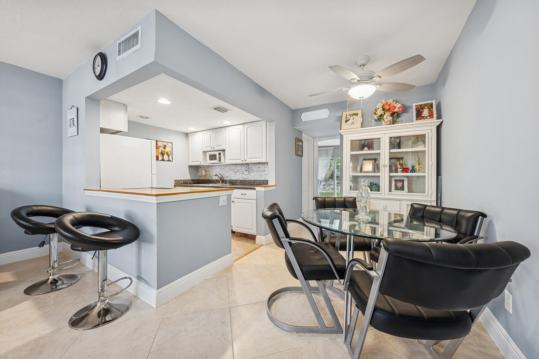Dining area with glass table and black chairs beside a kitchen with white cabinets and breakfast bar.
