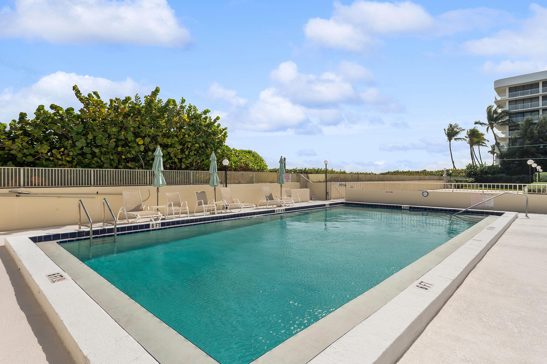 Condominium swimming pool bordered by lounge chairs, closed umbrellas, and tropical landscaping.