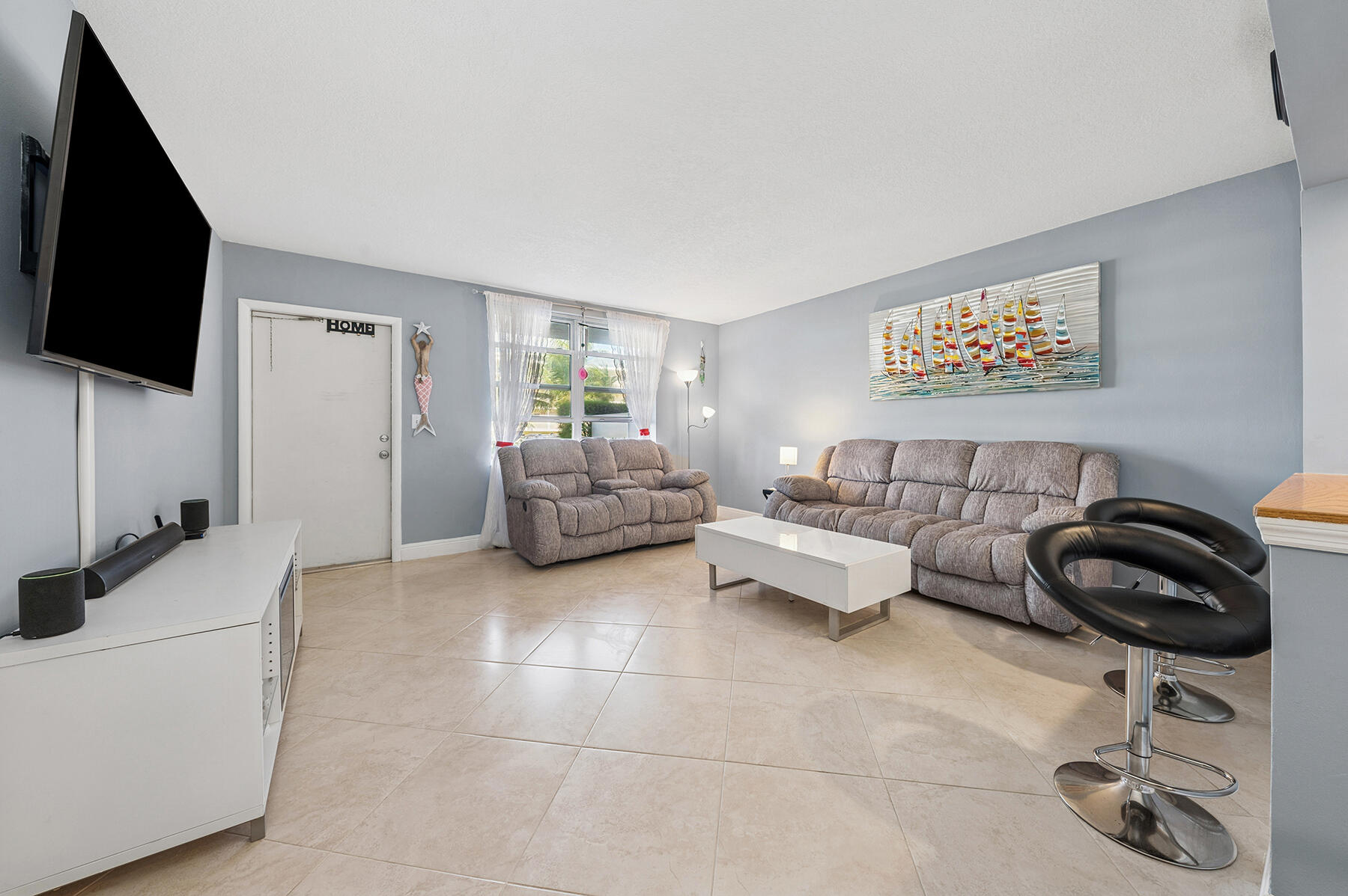 Furnished living room with gray reclining sofas, tile floors, and colorful sailboat artwork.