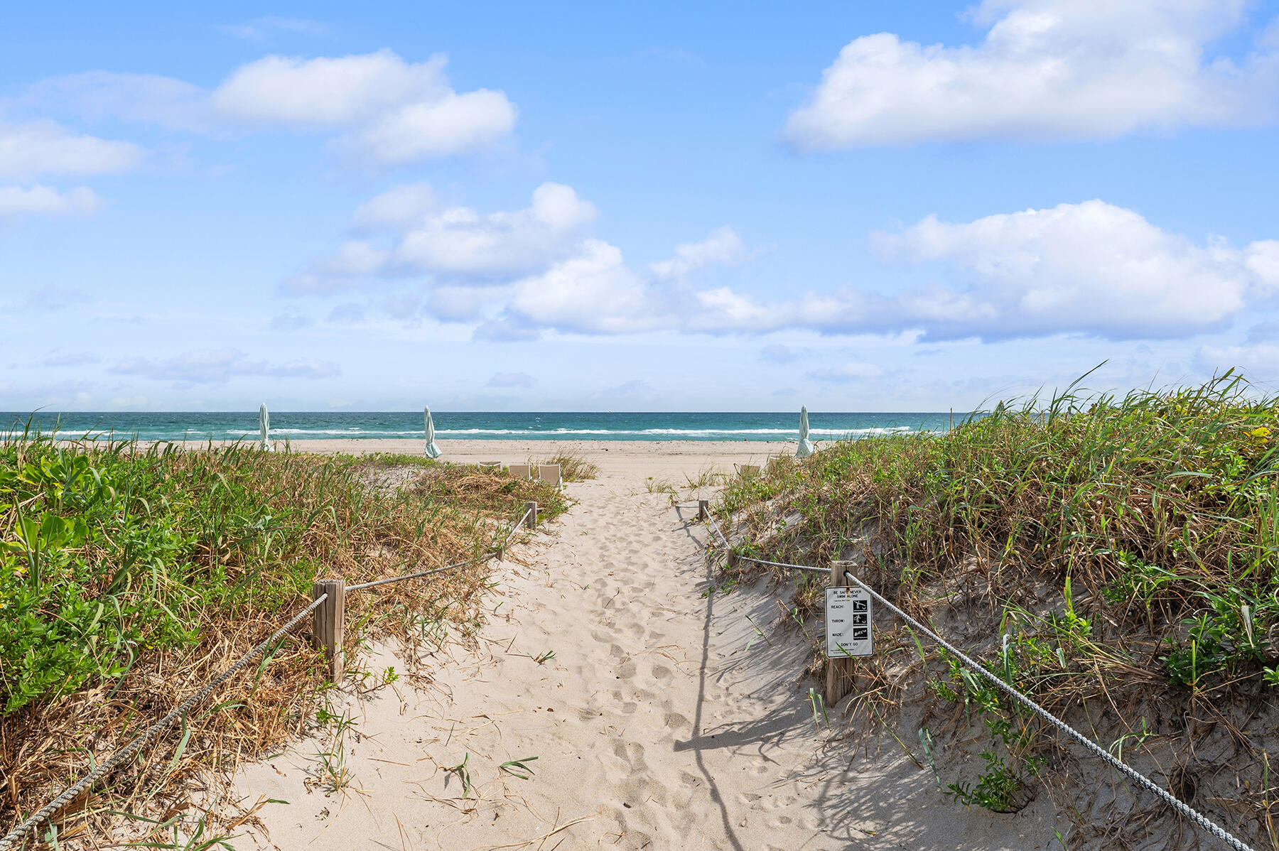 Sandy path through dune grasses leads to a beach with closed umbrellas and ocean views.