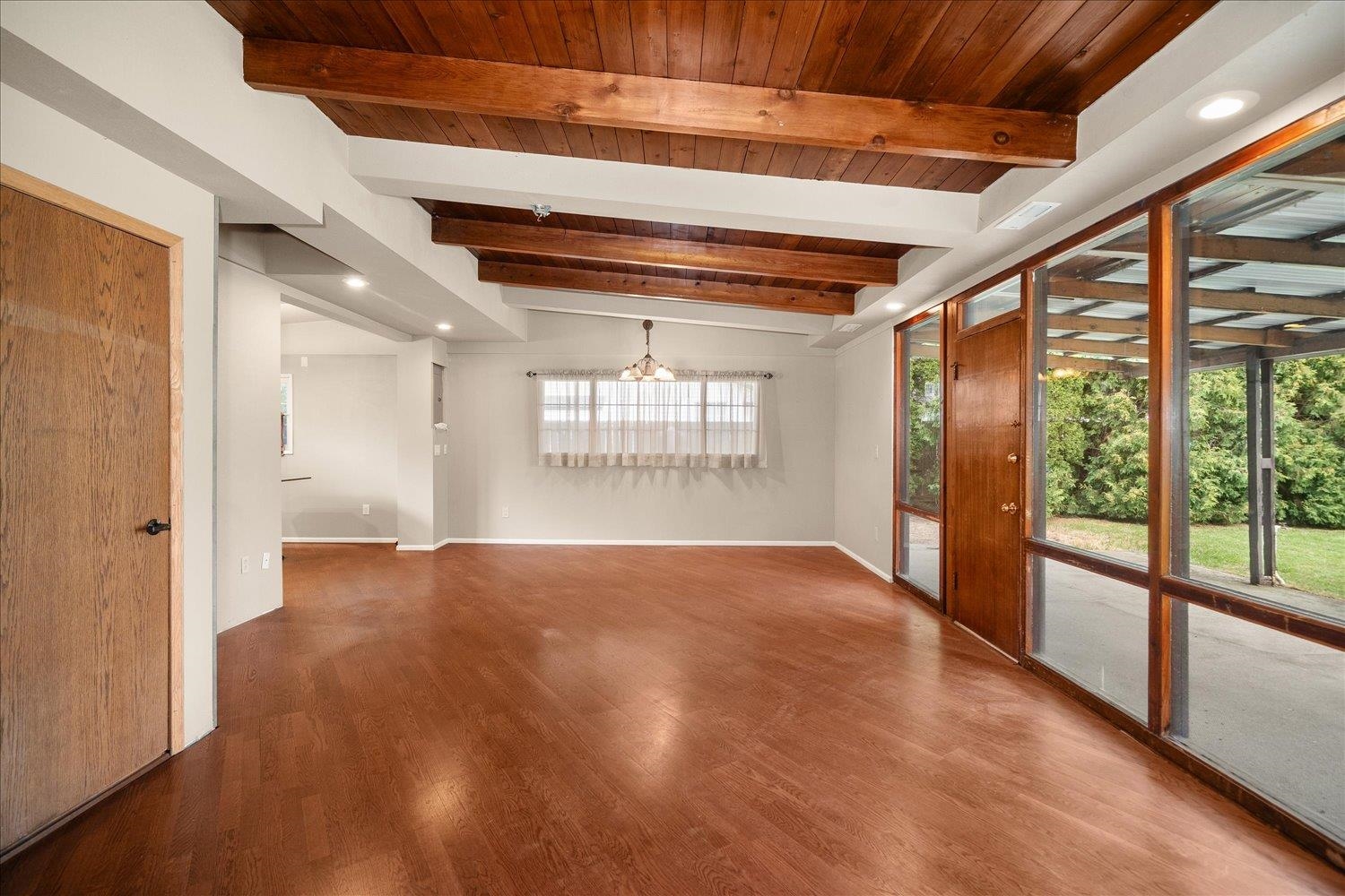 Empty living area with exposed wood ceiling beams, wood floors, and large windows facing a covered patio.