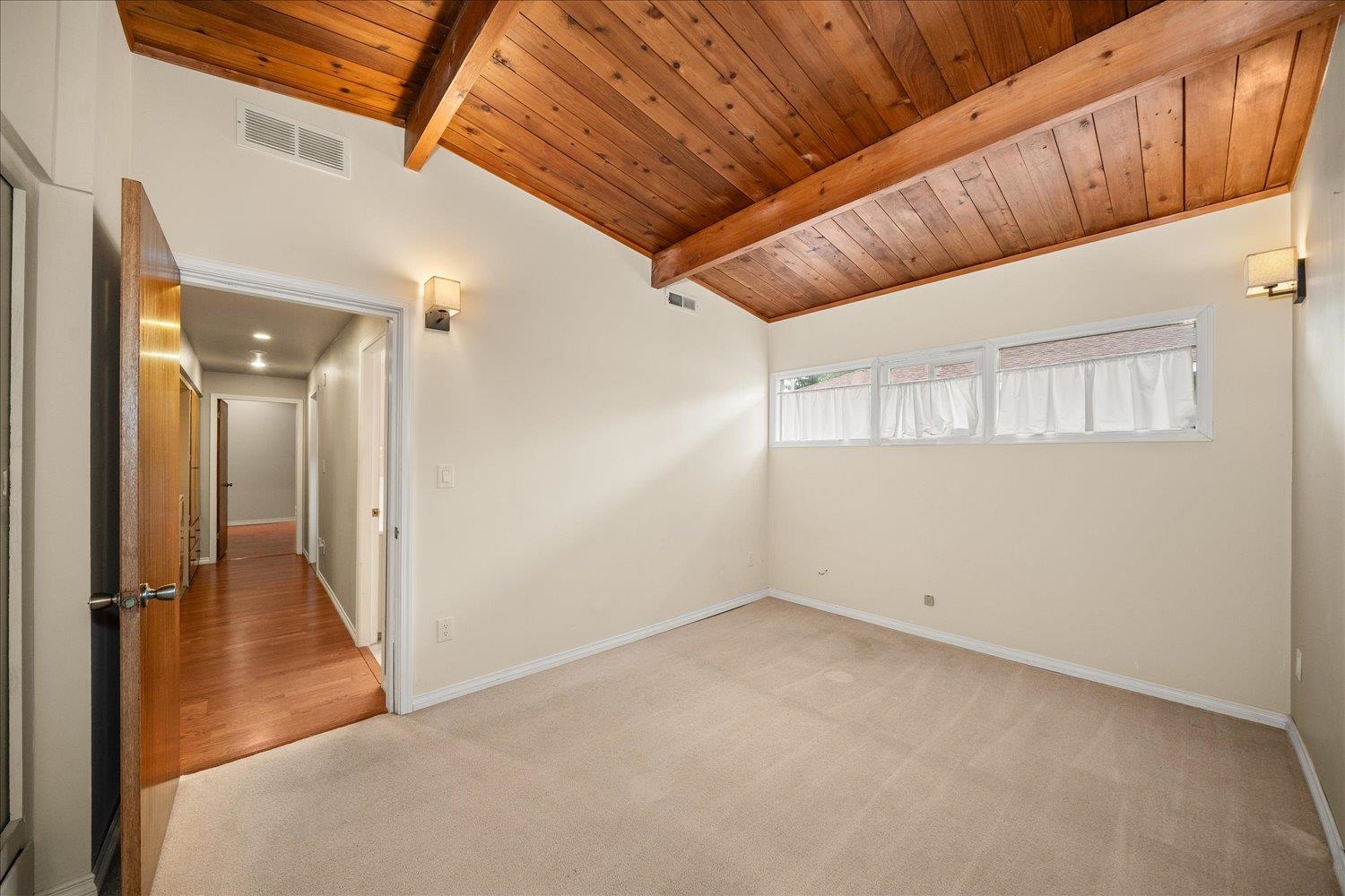 Empty bedroom with a vaulted wood ceiling, exposed beams, beige carpet, and narrow windows.