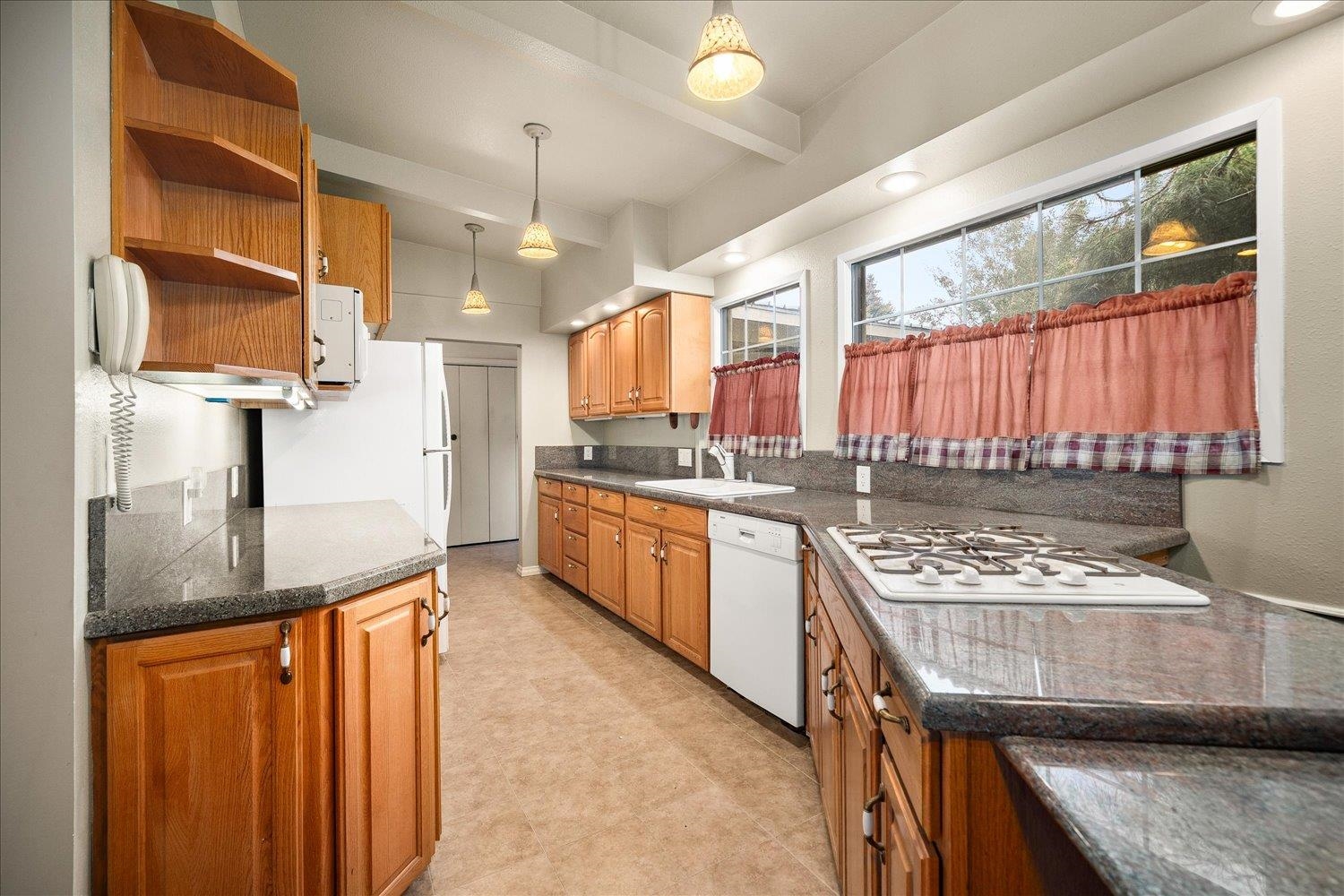 Long kitchen with warm wood cabinets, dark stone counters, gas cooktop, and large curtained windows.
