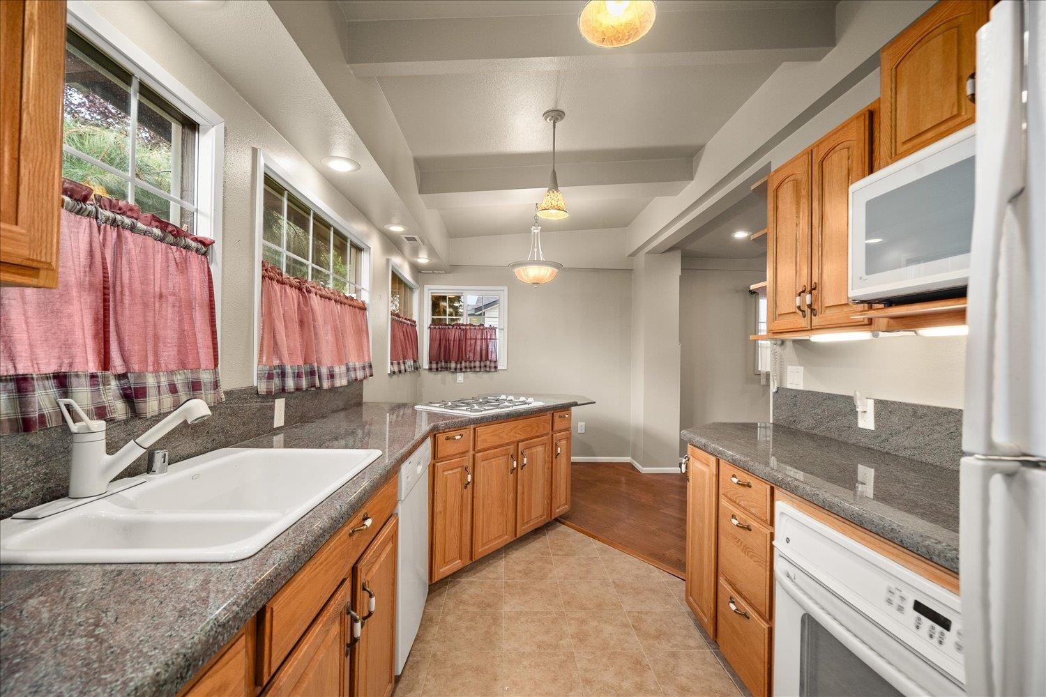Kitchen with wood cabinets, gray stone counters, a double-basin sink, and windows with pink curtains.